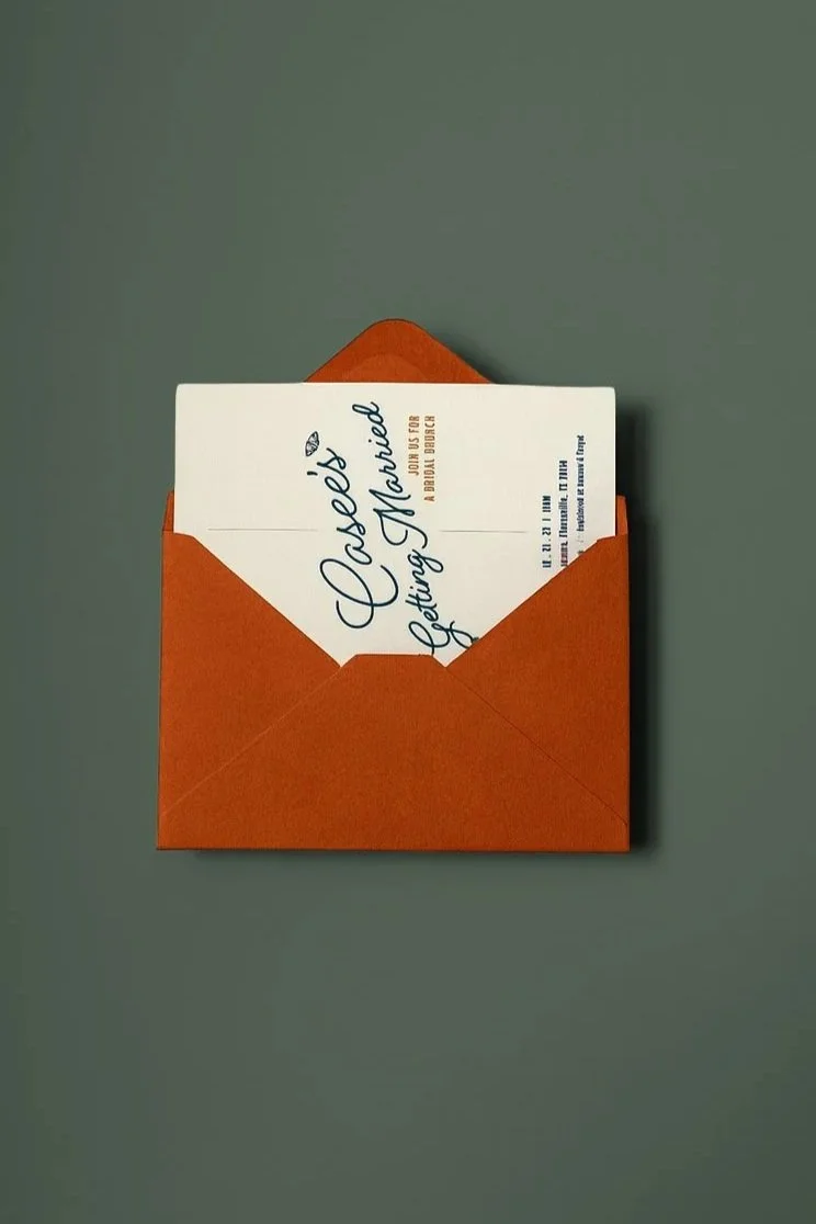 A wedding invitation card partially inside an orange envelope on a gray background.