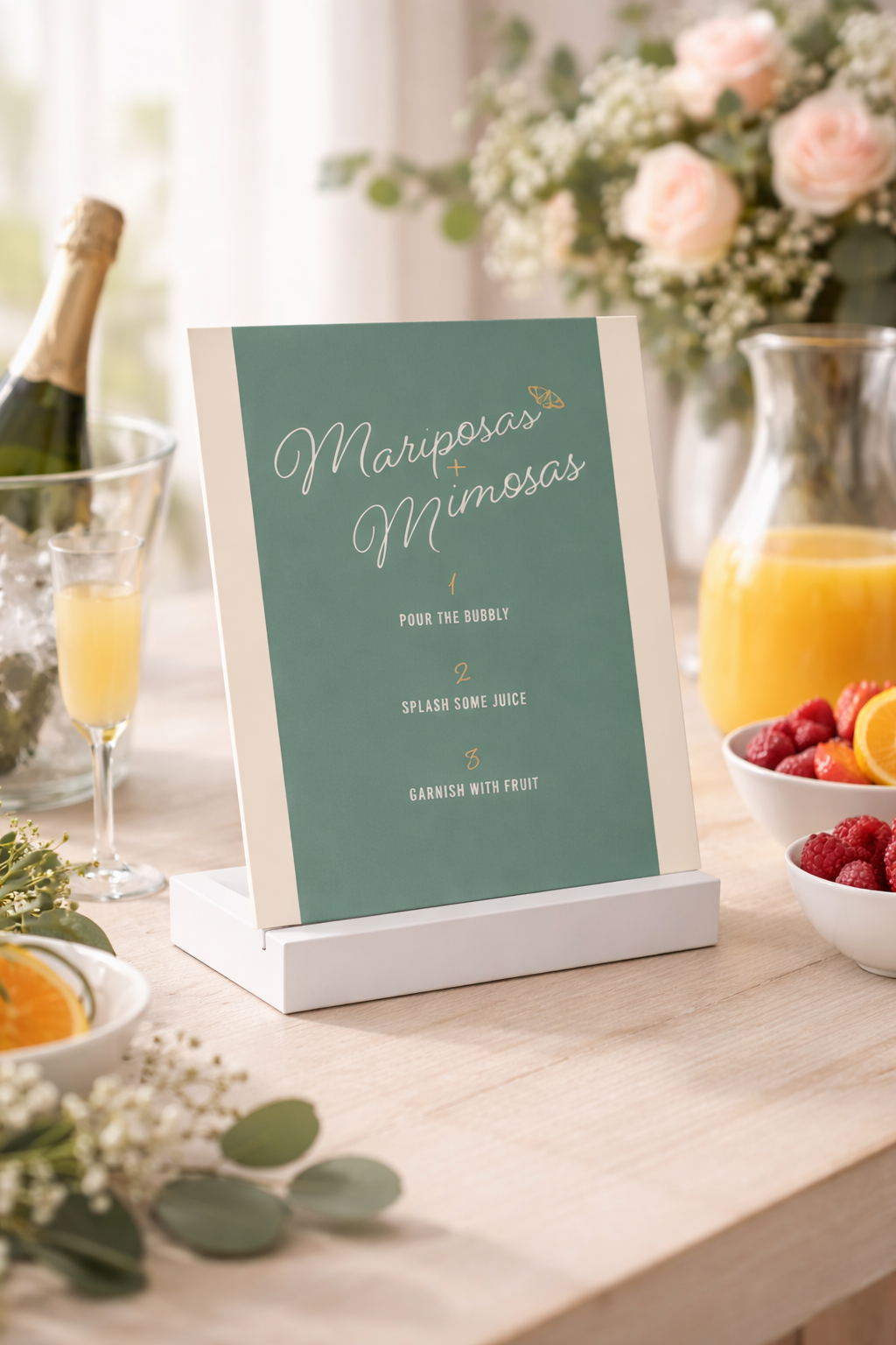 A welcome sign for a beverage station titled 'Mariposas + Mimosas' with instructions to pour the bubbly, splash some juice, and garnish with fruit, surrounded by bowls of fruit, a pitcher of orange juice, a glass of mimosa, a bottle of champagne in an ice bucket, and flower arrangements.
