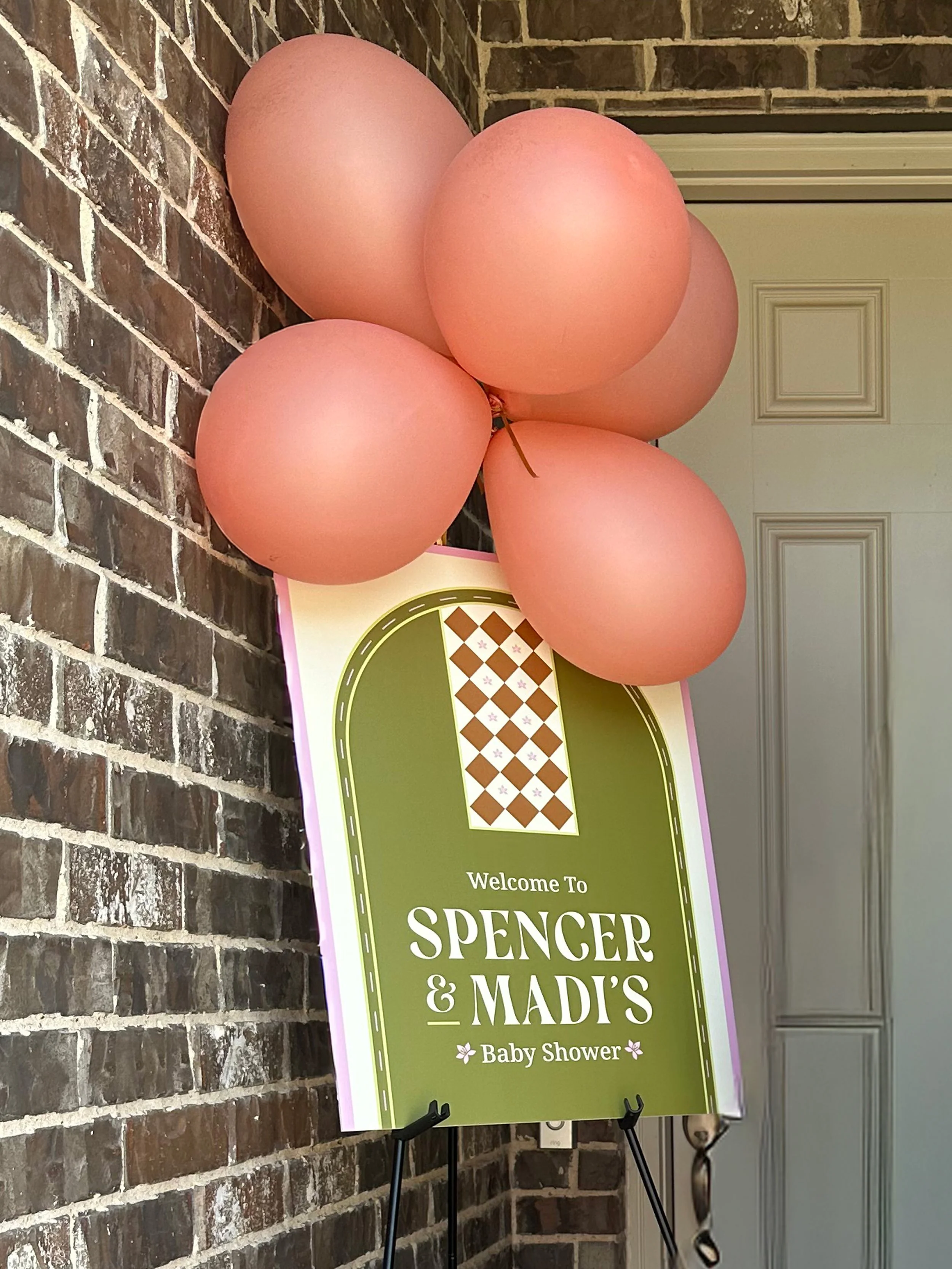 Baby shower sign with pink balloons attached to a brick wall, welcoming Spencer and Madi.