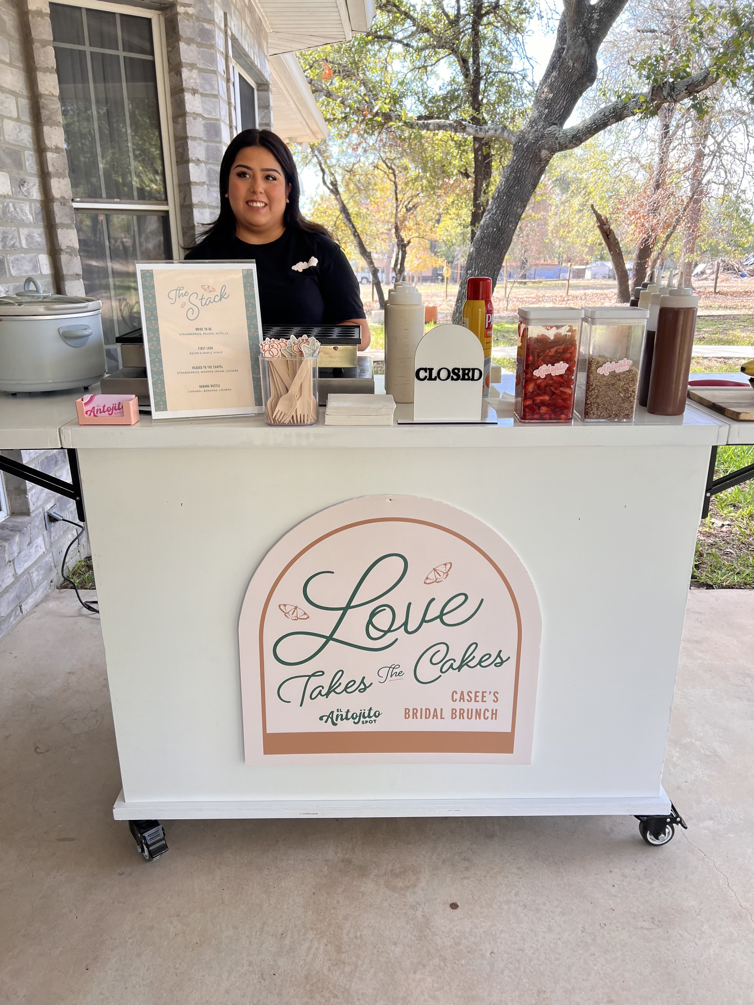 A woman stands behind a white cake cart with a sign that reads "Love Takes The Cakes" and "Casee's Bridal Brunch" while smiling. The cart has various toppings and condiment bottles on top, with trees and a fence visible in the background.