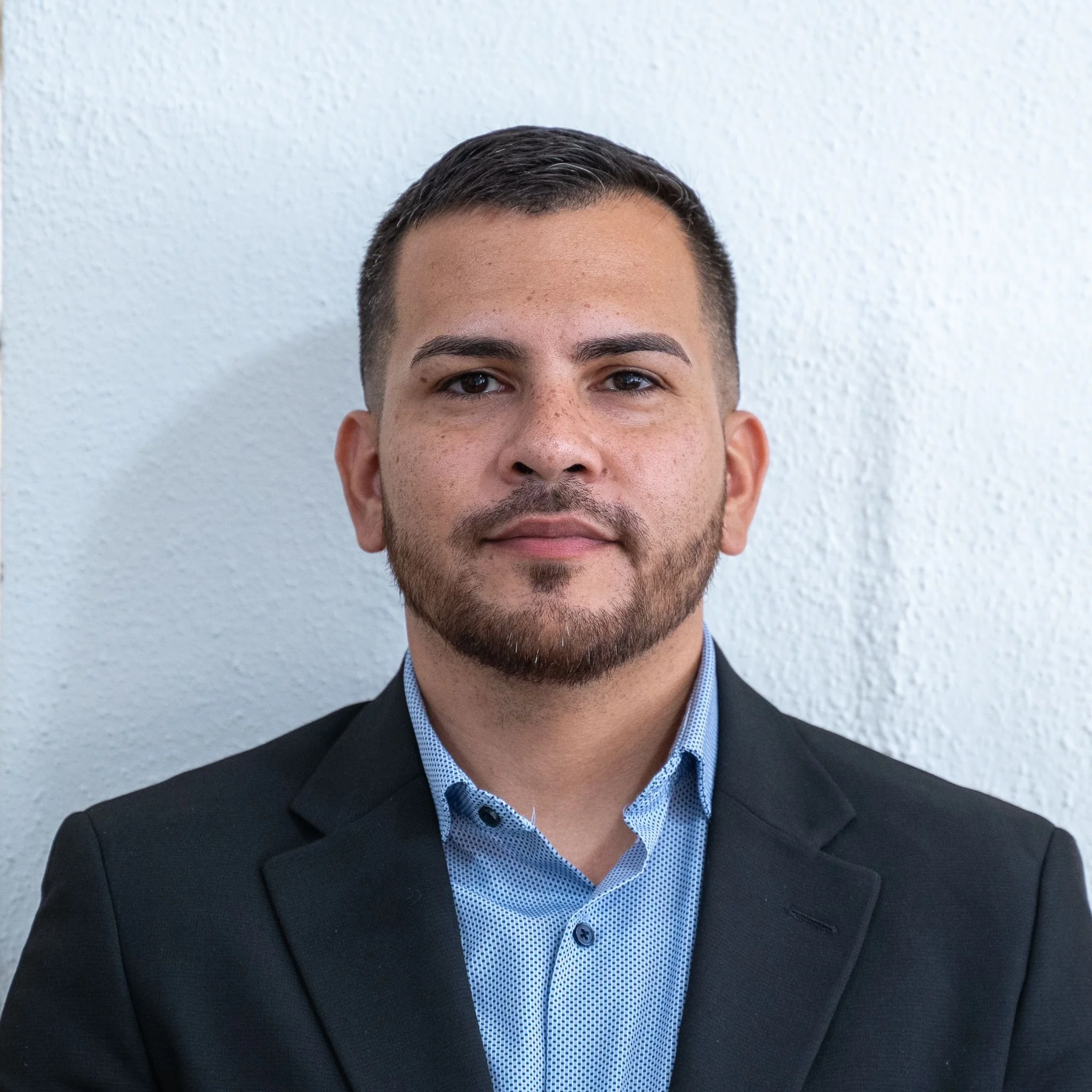 A man with short hair and a beard wearing a dark suit jacket and a light blue, button-up shirt, standing against a white textured wall.