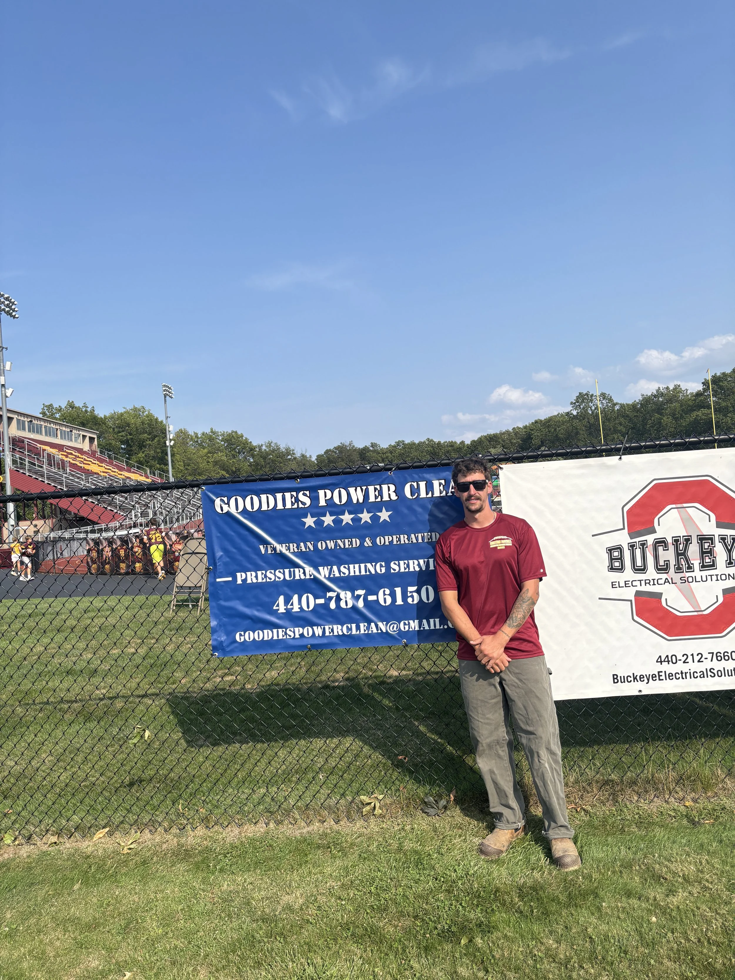 A man wearing sunglasses, a maroon t-shirt, and tan work boots standing in front of a chain-link fence with banners at a sports field. Behind him, there are spectators, some in sports uniforms, and a bleacher section under a clear blue sky.