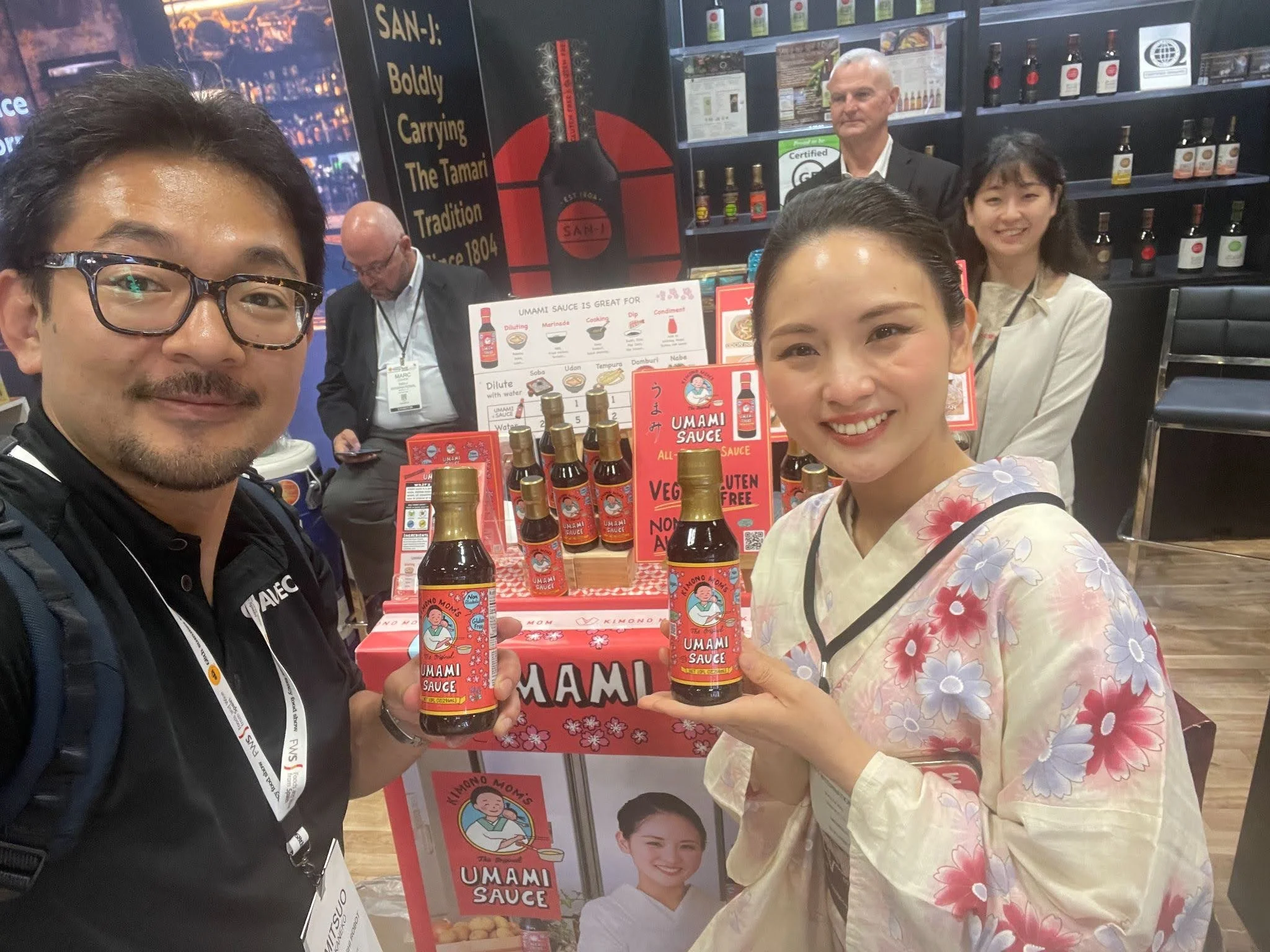 Two people, a man and a woman, holding bottles of Umami Sauce at a booth promoting the product. In the background, there are other people, display shelves with bottles, and promotional signs for the sauce.
