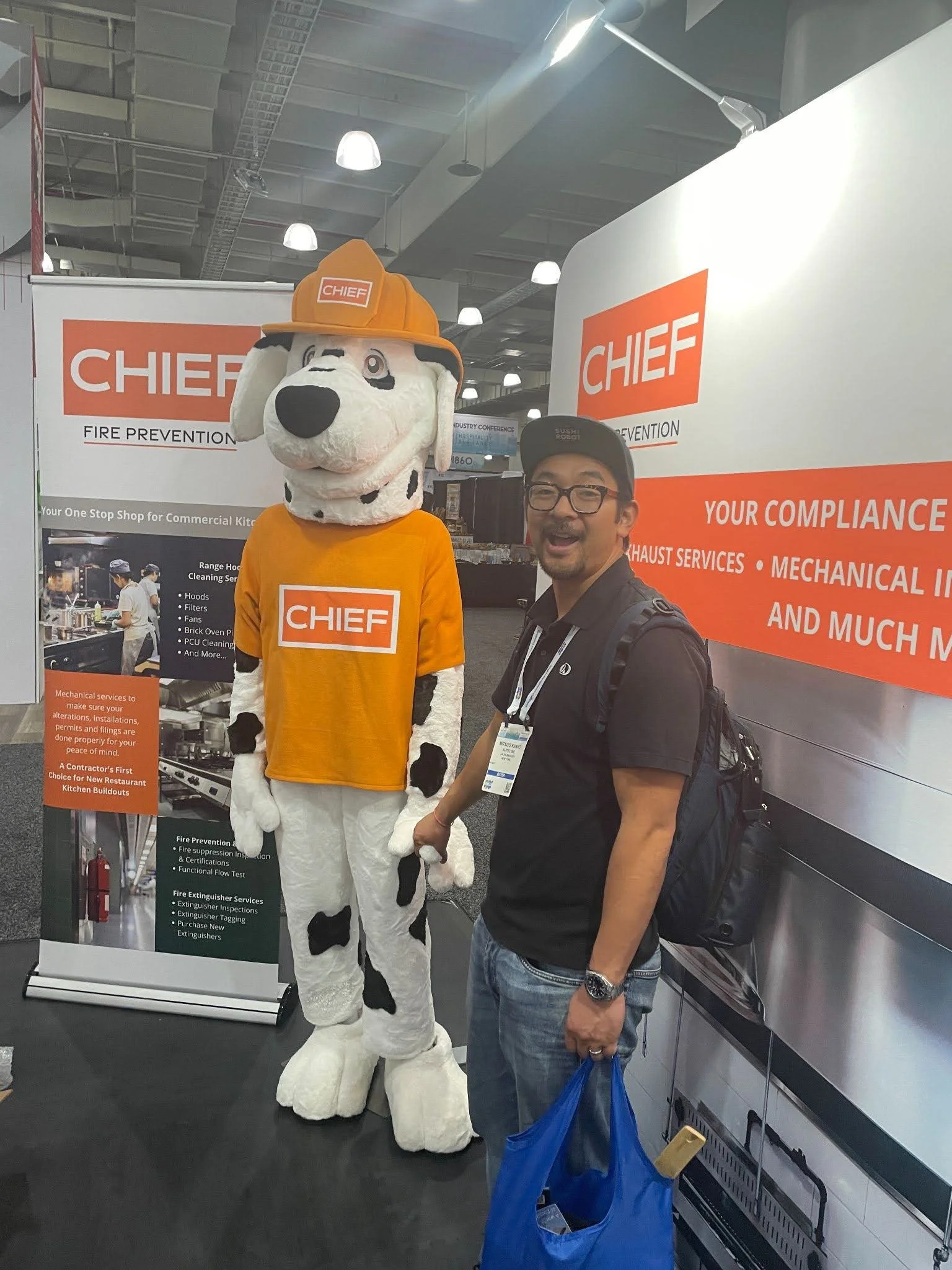 A man standing next to a mascot dressed as a Dalmatian dog wearing a fire chief helmet and an orange t-shirt, both at a trade show booth for a company called CHIEF, which specializes in fire prevention and mechanical services.