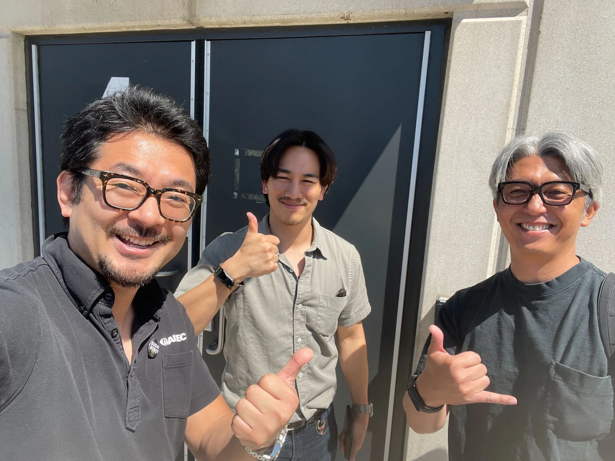Three smiling men standing outdoors in front of a dark door, giving thumbs-up and hand gestures.
