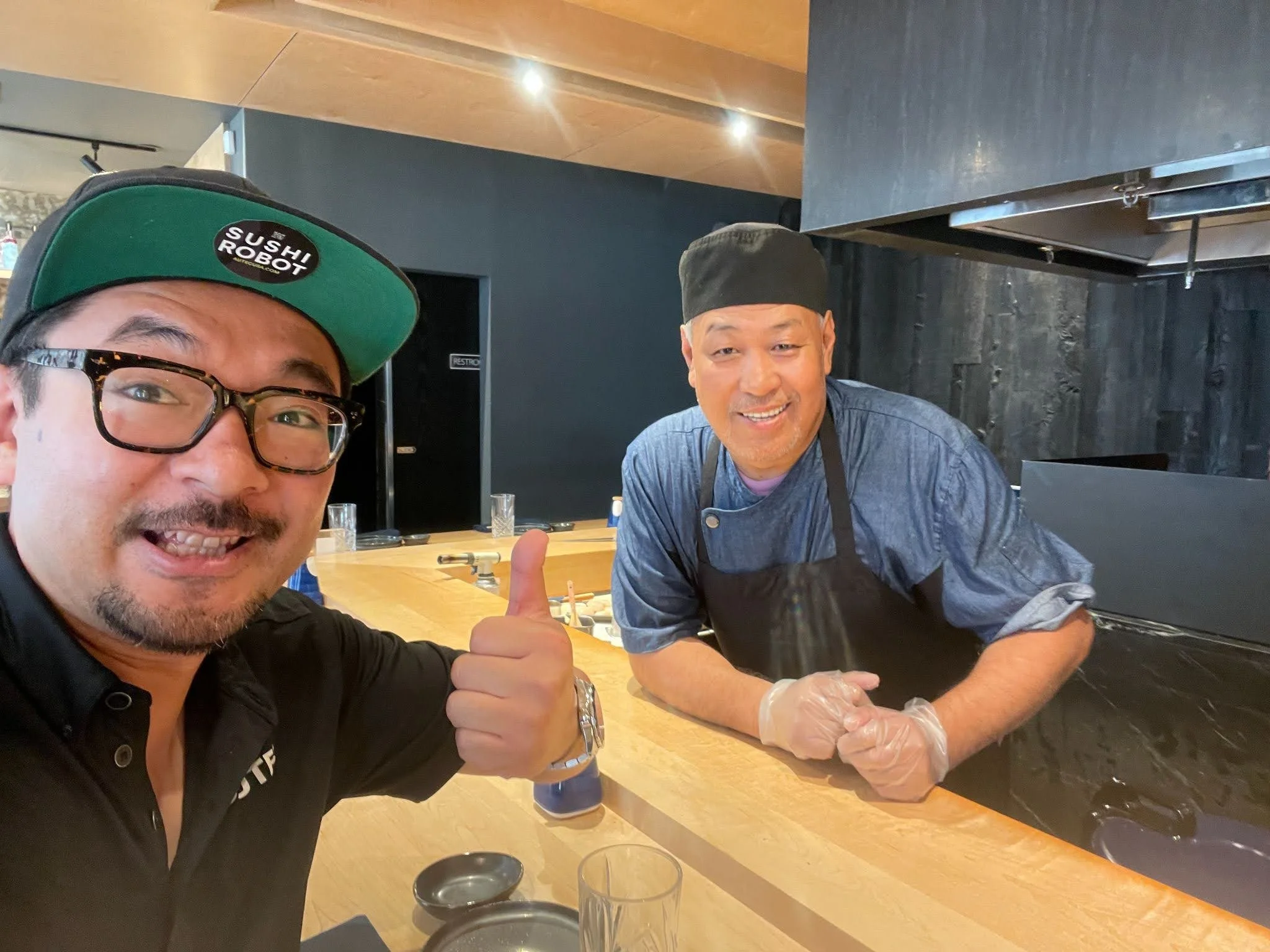 Two men in a sushi restaurant. One man is wearing glasses and a black cap that says sushi robot, giving a thumbs up. The other man is a sushi chef wearing a black hat and apron, smiling and leaning on the wooden counter.
