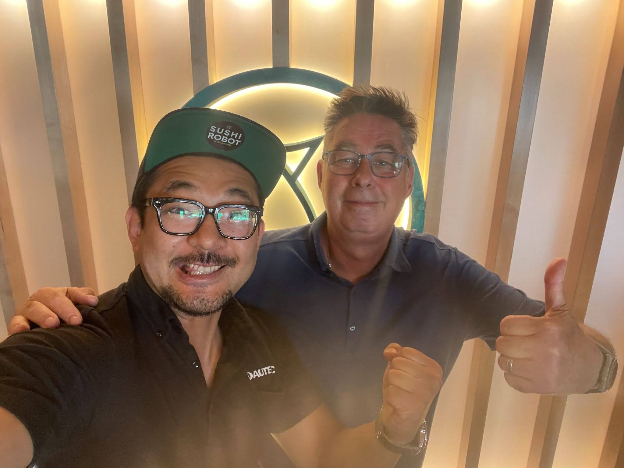 Two men smiling and posing for a selfie indoors, one wearing glasses, a black shirt with a 'DANTE' label and a green cap that says 'SUSHI ROBOT', the other giving a thumbs-up, both standing in front of wooden paneling with a glowing circular logo in 