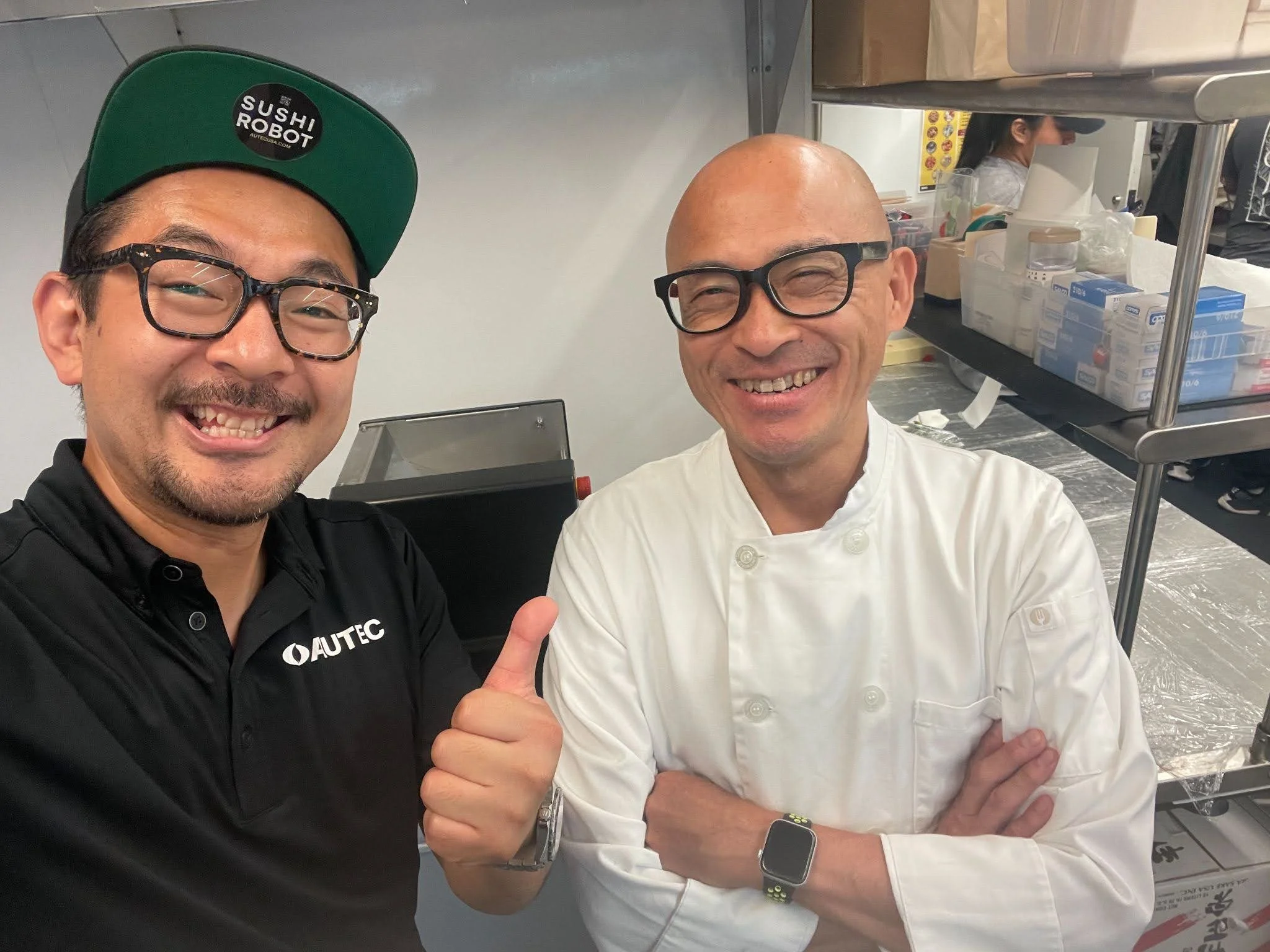 Two men smiling and taking a selfie in a kitchen, one wearing glasses and a cap with a sushi robot logo, and the other is a chef in a white uniform with crossed arms.