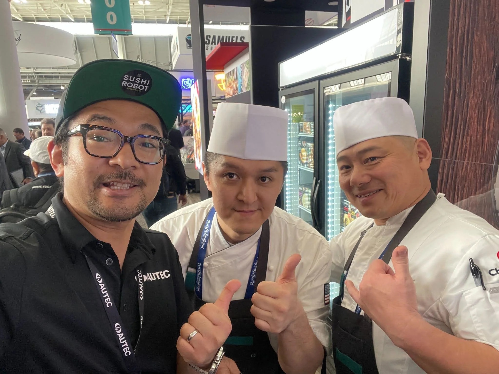 Three men in chef uniforms and a man in casual clothing posing together at a trade show or event, all giving thumb-up signs.