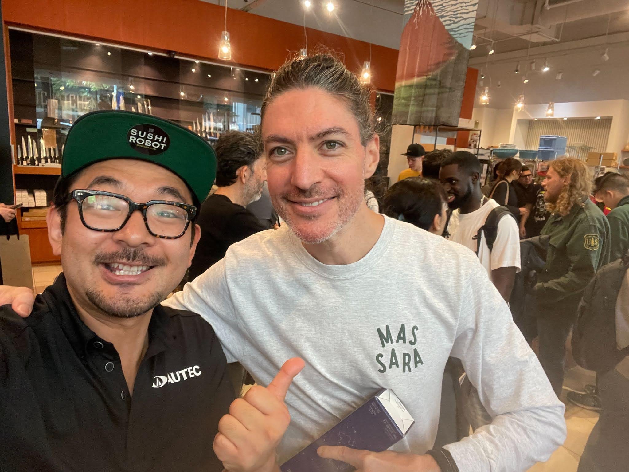 Two men smiling at a busy indoor event, one wearing glasses, a black polo shirt with a logo, and a green cap that says 'SUSHI ROBOT'; the other wearing a light grey long-sleeve shirt and holding a boxed product, with a crowd of people in the backgrou