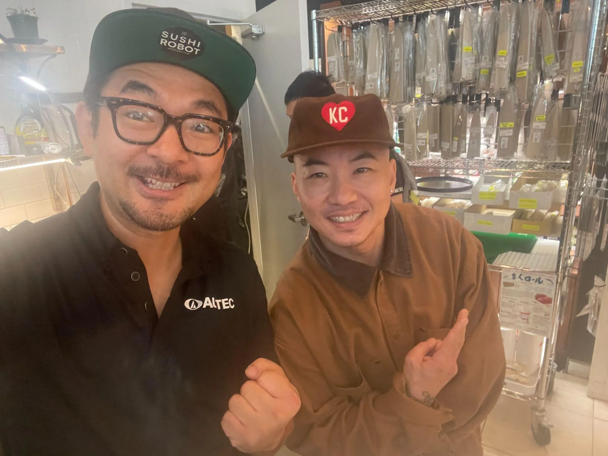 Two smiling men taking a selfie in a kitchen or store, with shelves holding knives and kitchen supplies behind them. One man is wearing glasses, a black polo with 'ALTEC' on it, and a black cap that says 'SUSHI ROBOT.' The other person is pointing at