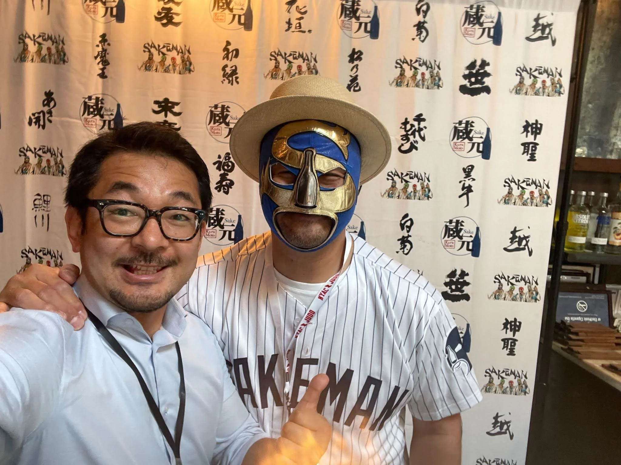 Two men posing for a photo, one wearing glasses and a white shirt, the other wearing a luchador mask, white fedora, and a cricket jersey with "SAKEMAN" on it, standing in front of a backdrop with Japanese characters and the word "SAKEMAN" repeated.