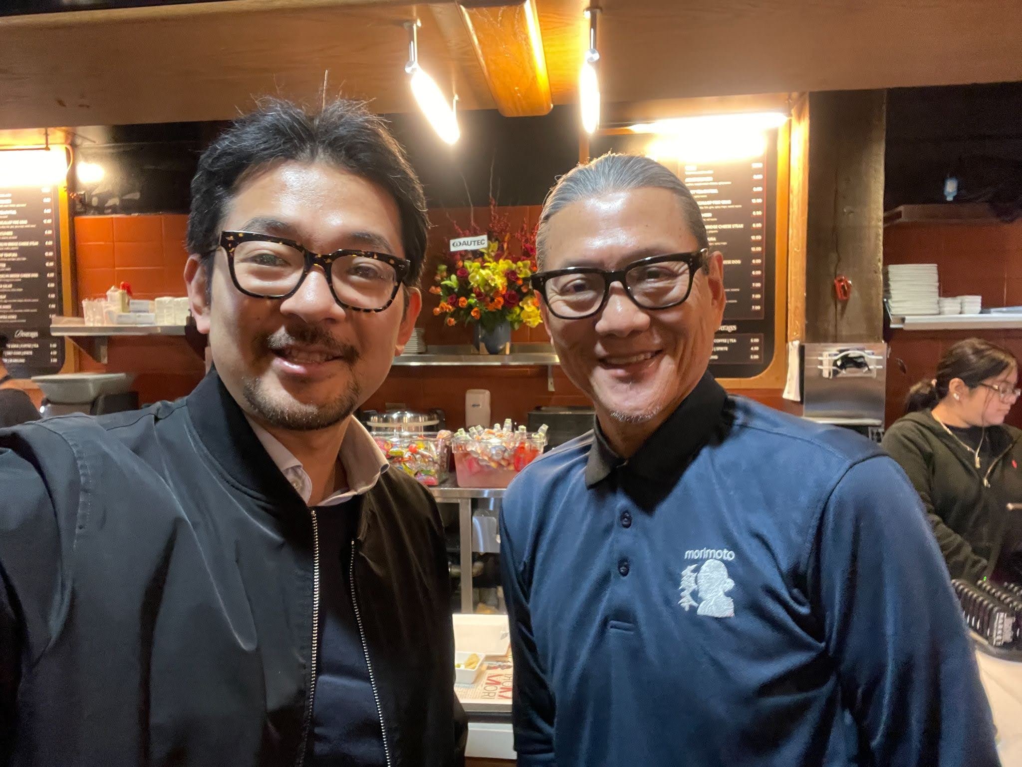 Two men smiling at a camera, standing in an indoor setting with a menu board and flowers in the background.