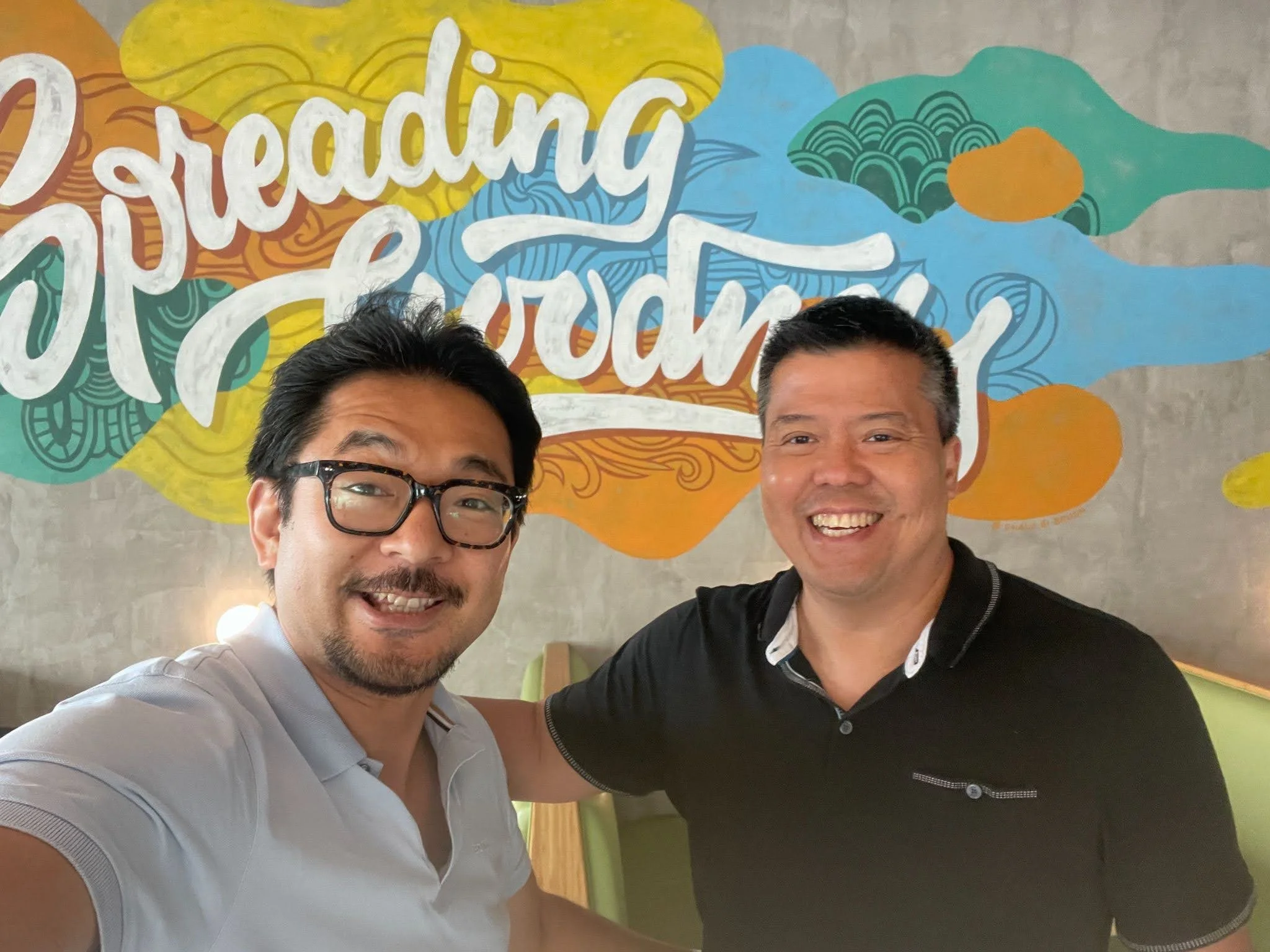Two men smiling inside a restaurant with a colorful mural behind them that says 'Sharing a Product'.