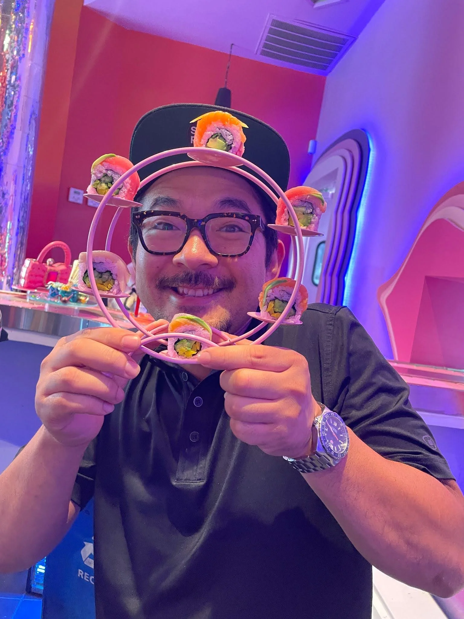 Man smiling and holding a pink circular stand with sushi rolls arranged around it in a colorful, neon-lit room.