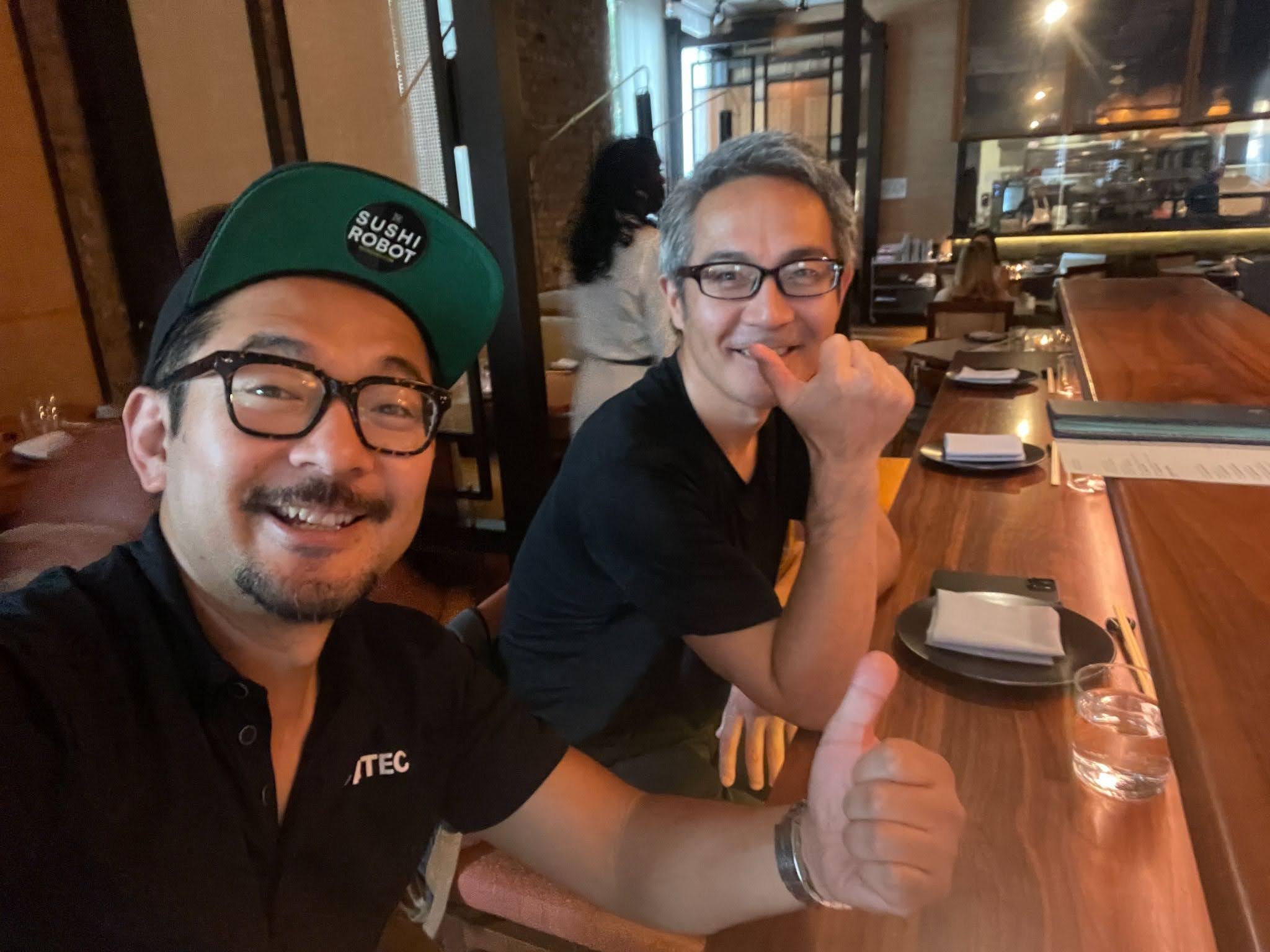 Two men sitting at a restaurant table, smiling at the camera. One wearing glasses and a green cap with 'SUSHI ROBOT' badge, the other resting his chin on his hand, giving a thumbs-up. The table has menus, plates, napkins, and a glass of water.