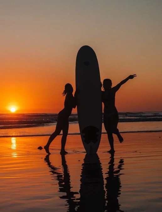 Silhouettes of two women with a surfboard on the beach during sunset.