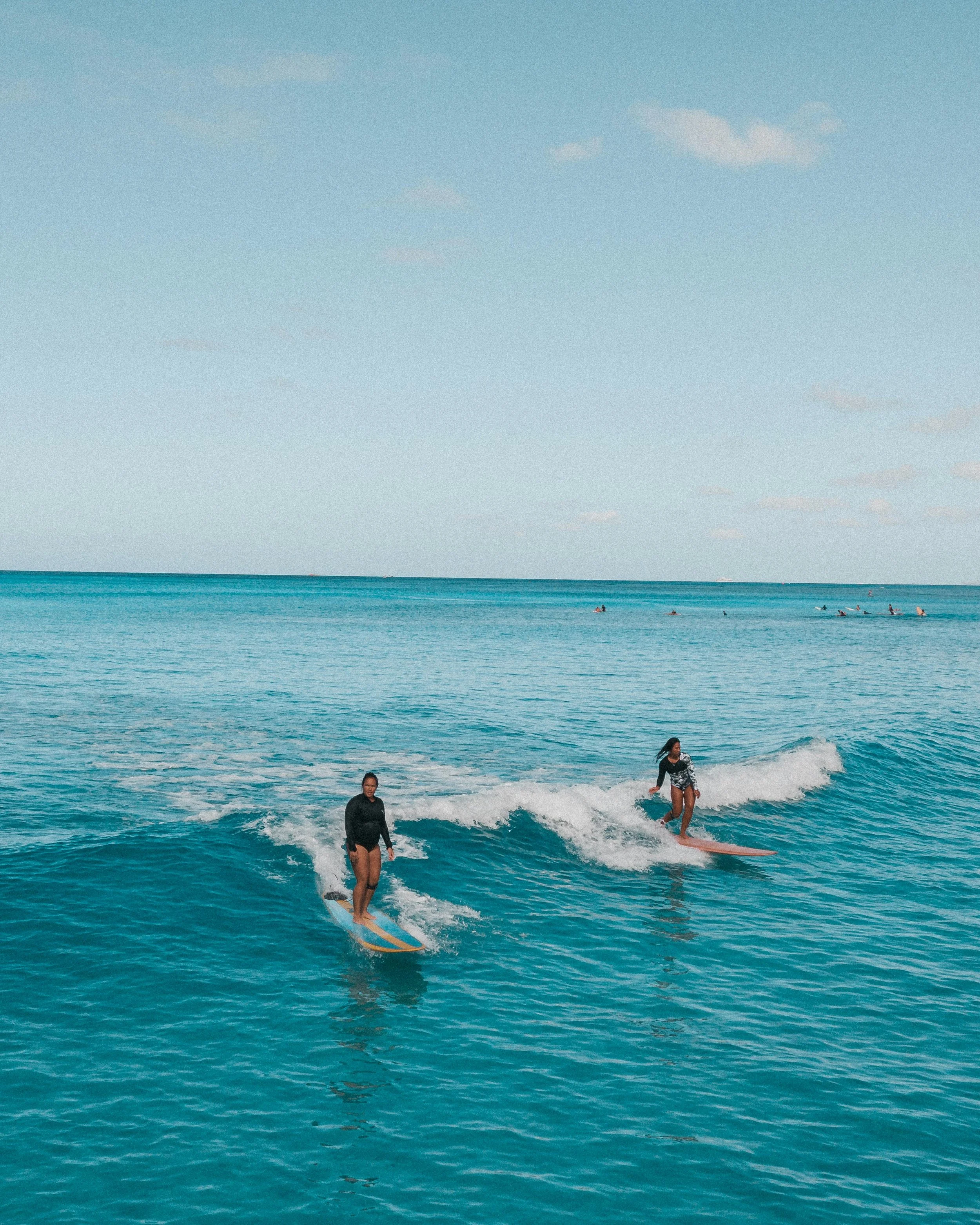 Two women surfing on the ocean, one in black attire and the other in a black and white wetsuit, with a clear blue sky and distant surfers in the background.