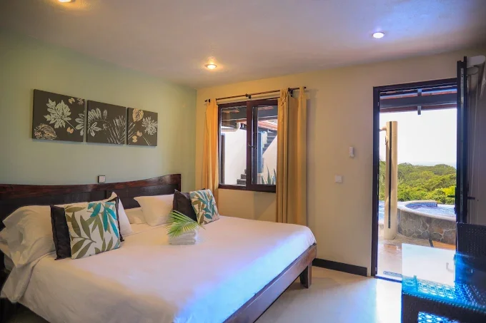 Hotel room with a large bed, decorative pillows, a window with yellow curtains, and an open door leading outside to a balcony with a hot tub and a view of green trees.