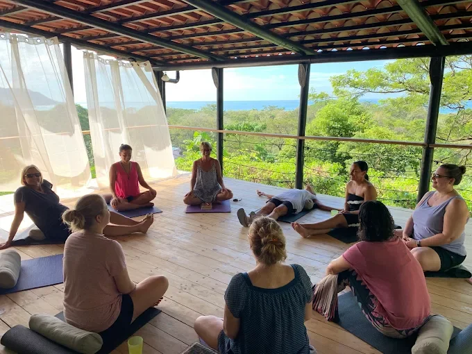 Group of women practicing yoga or meditation on mats inside a wooden-framed pavilion with large open sides, overlooking lush green trees and a distant view of the ocean under a partly cloudy sky.