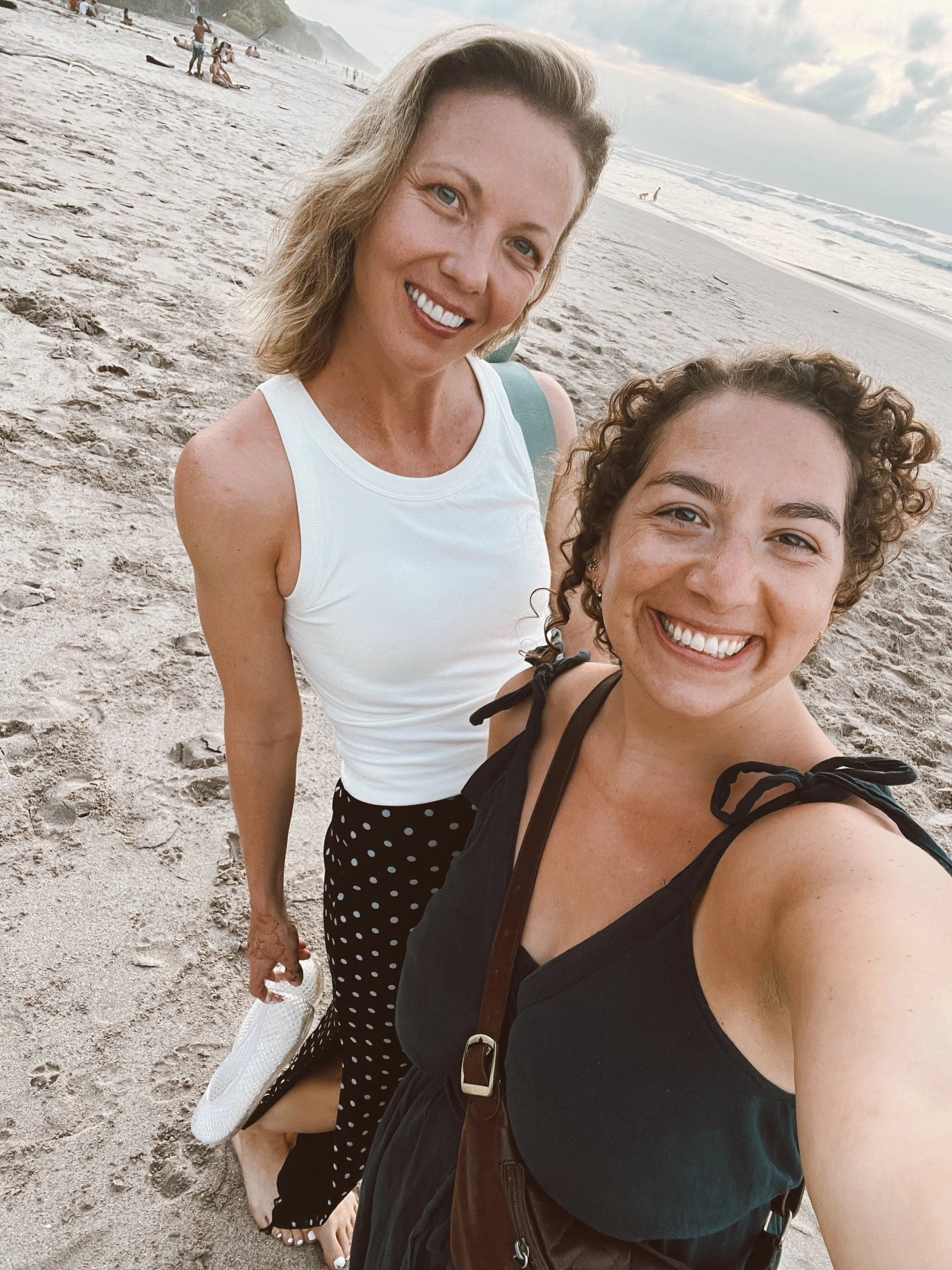 Two women smiling on a beach, taking a selfie with the ocean and other beachgoers in the background.