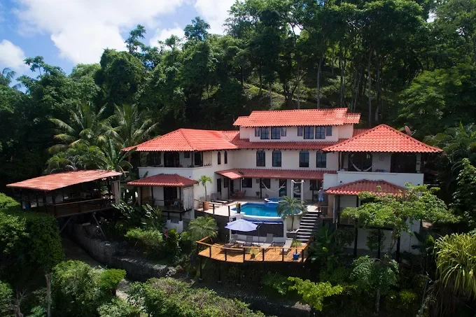 Large white house with multiple red-tiled roofs, surrounded by trees, with a swimming pool and deck area in the backyard.