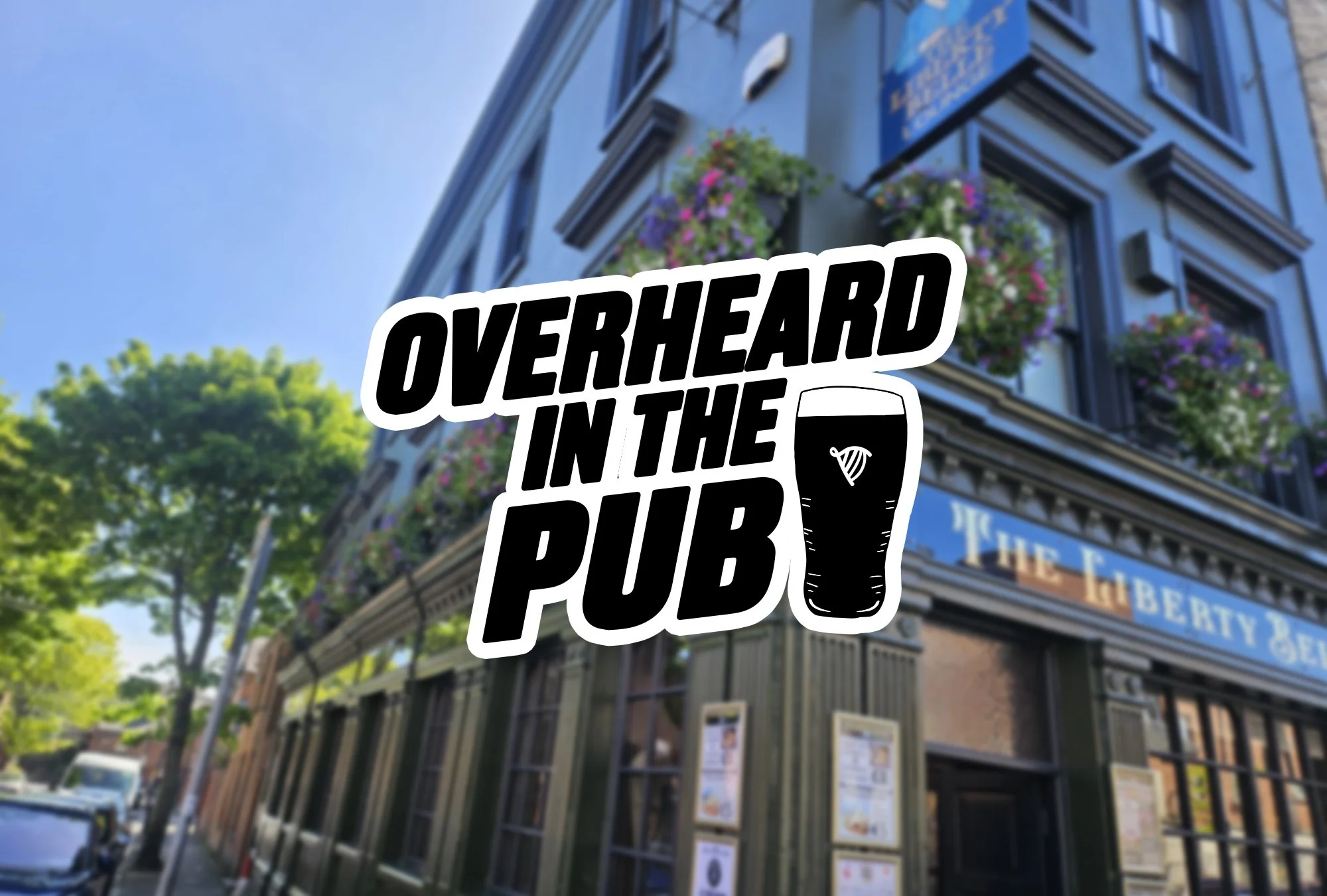Sign with the text "Overheard in the Pub" and a pint of beer illustration in front of a pub building with blue exterior, flower baskets, and green trees.