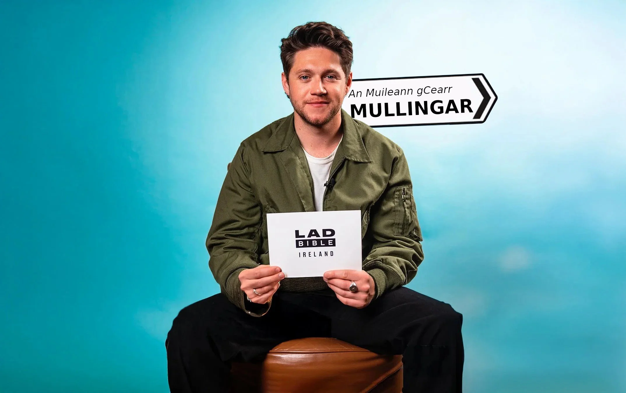 A man sitting on a stool against a blue background, holding a card with the text "LAD BIBLE IRELAND." There is a sign behind him with the text "An Muileann gCearr MULLINGAR." He is wearing a green jacket, a white shirt, and black pants.