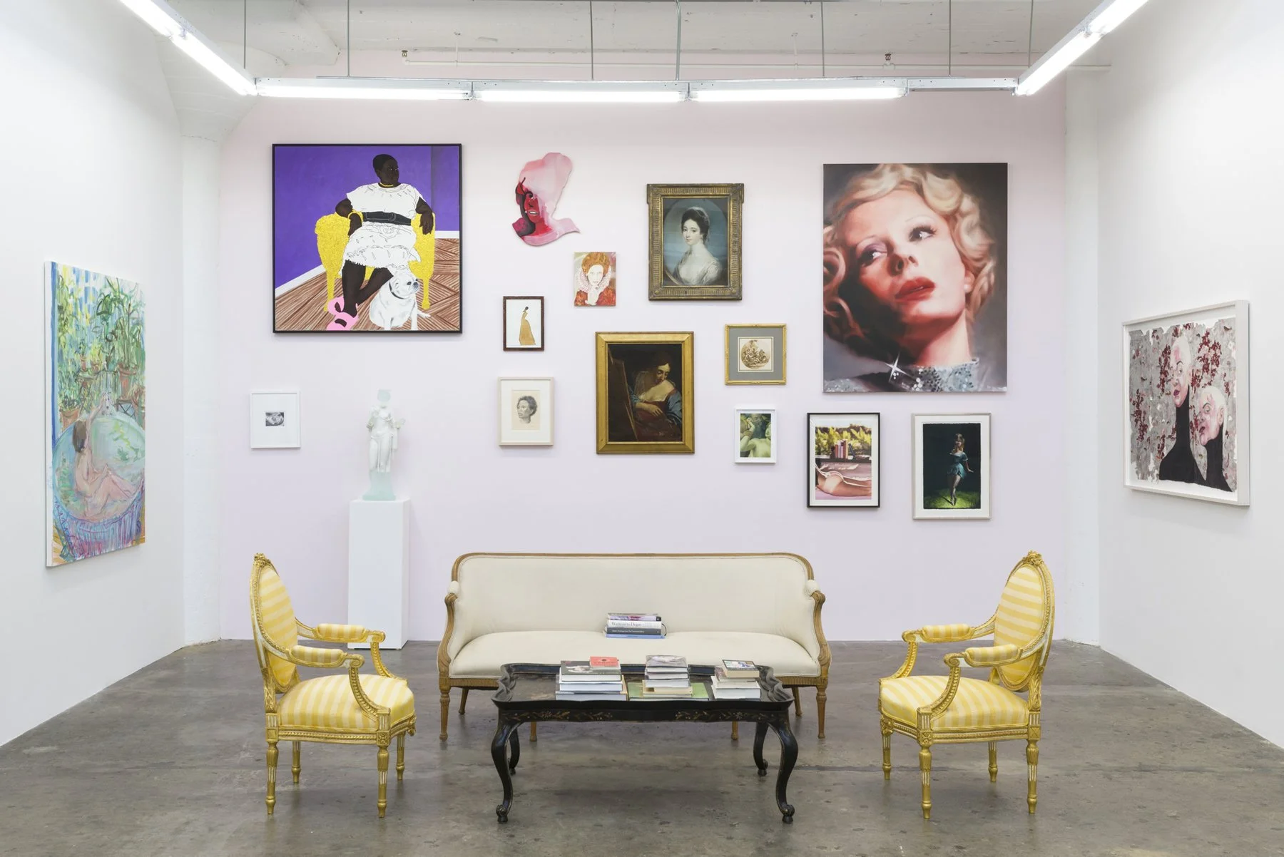 An art gallery with framed paintings and photographs on white walls, including a large portrait of a woman with curly blonde hair, a modern portrait of a woman with dark skin on a yellow chair, and other colorful artworks. There is a beige vintage sofa, two yellow striped armchairs, and a black coffee table with books in front of the seating area.