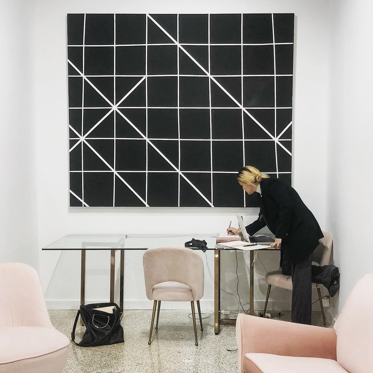 A woman dressed in black, leaning over a glass table, working on a laptop in a modern, minimalist office space with pink chairs and a large abstract black and white artwork on the wall.