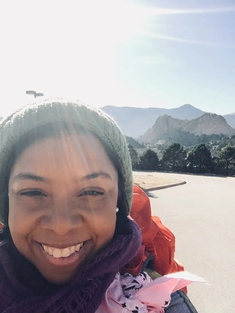 A woman smiling outdoors with mountains and a clear sky in the background, wearing a beanie and a backpack.