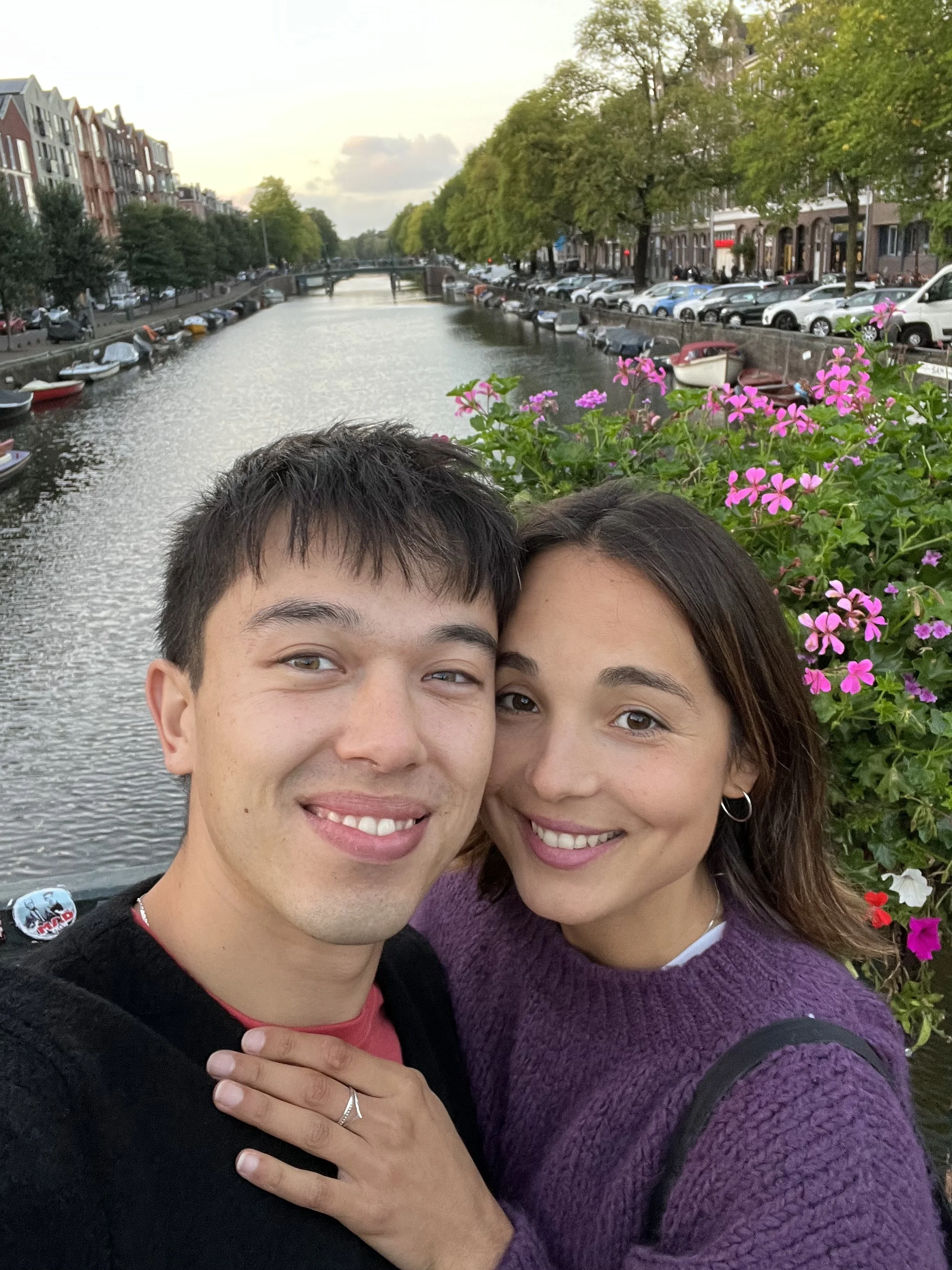 Un couple souriant pose près d'un canal bordé d'arbres et de bâtiments, avec des fleurs rose et violet en premier plan.
