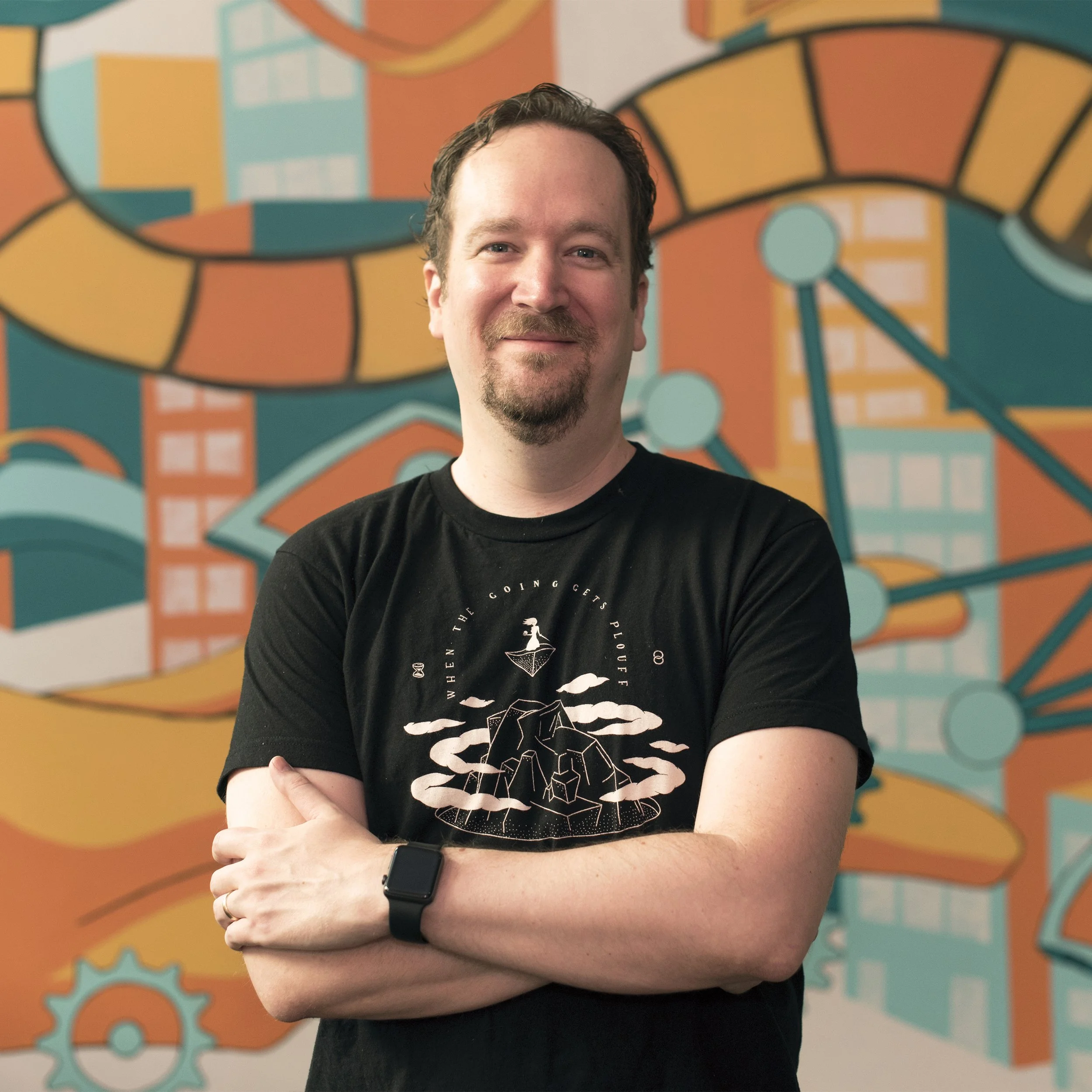 A man with a beard and mustache, smiling, standing with arms crossed in front of a colorful mural featuring geometric shapes and architectural elements.