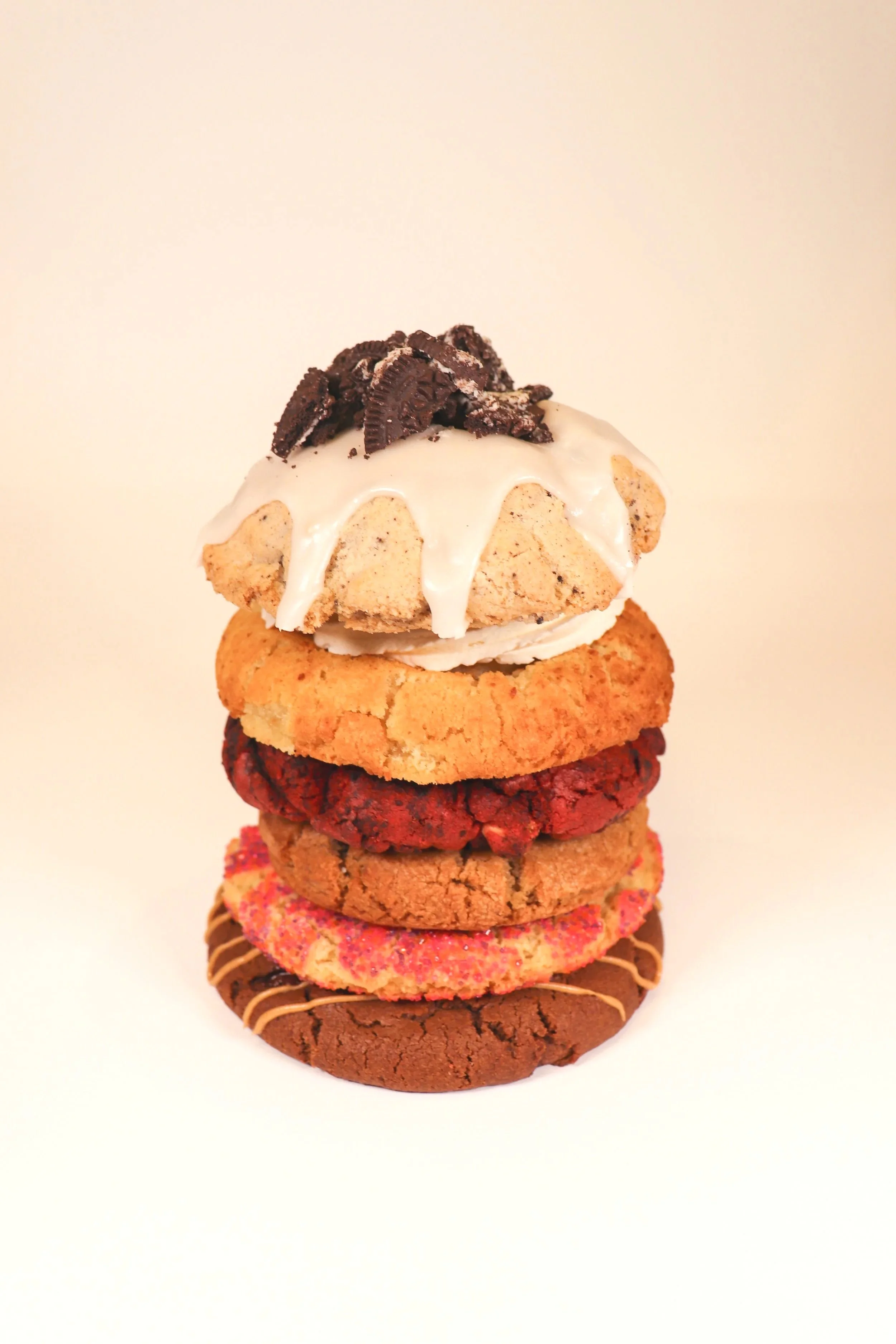 Stack of six assorted cookies with various toppings, including white icing and chocolate pieces on top, against a plain light background.