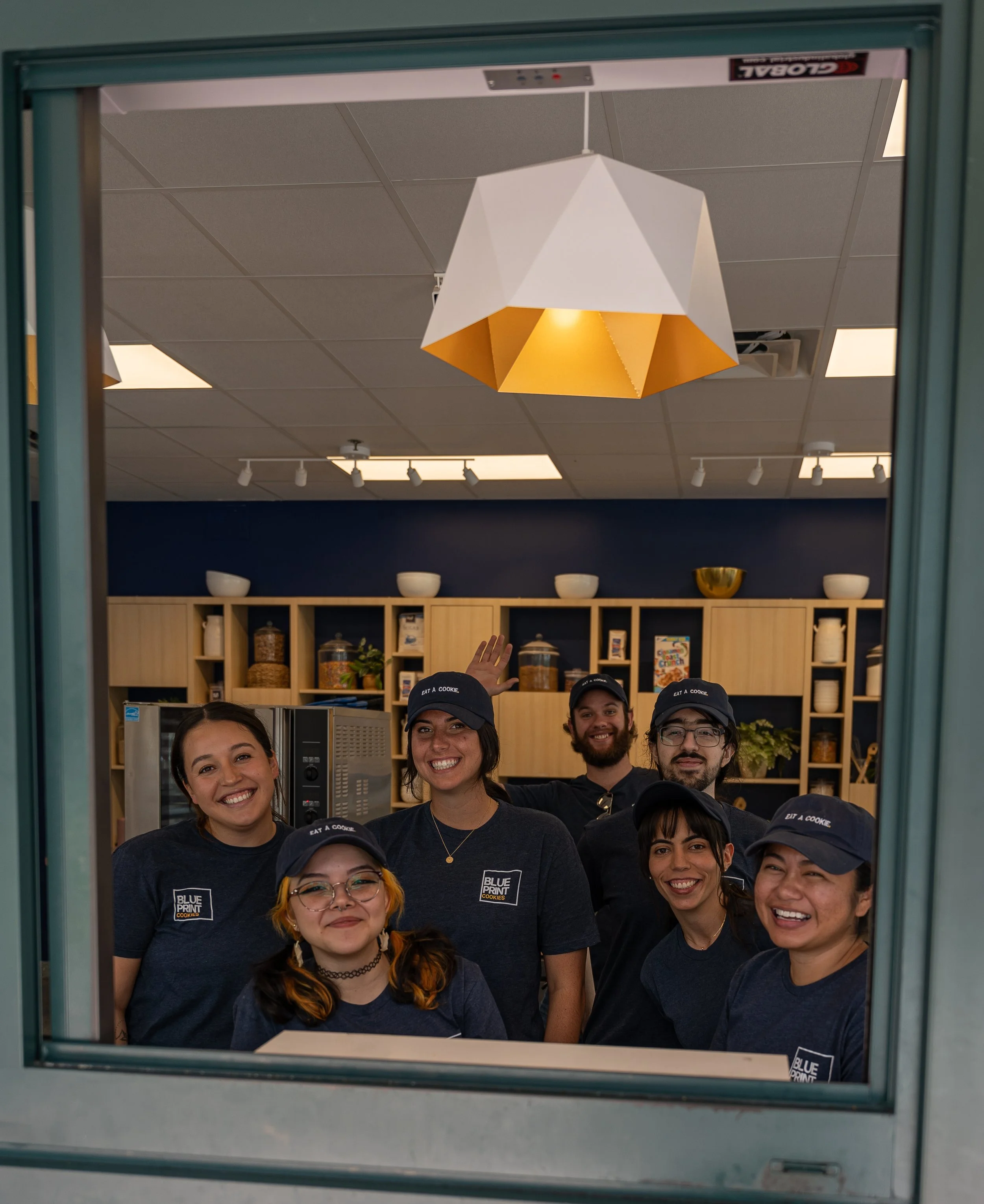 Group of six happy smiling people wearing dark blue t-shirts and caps, standing inside a kitchen or cafeteria, viewed through a service window.