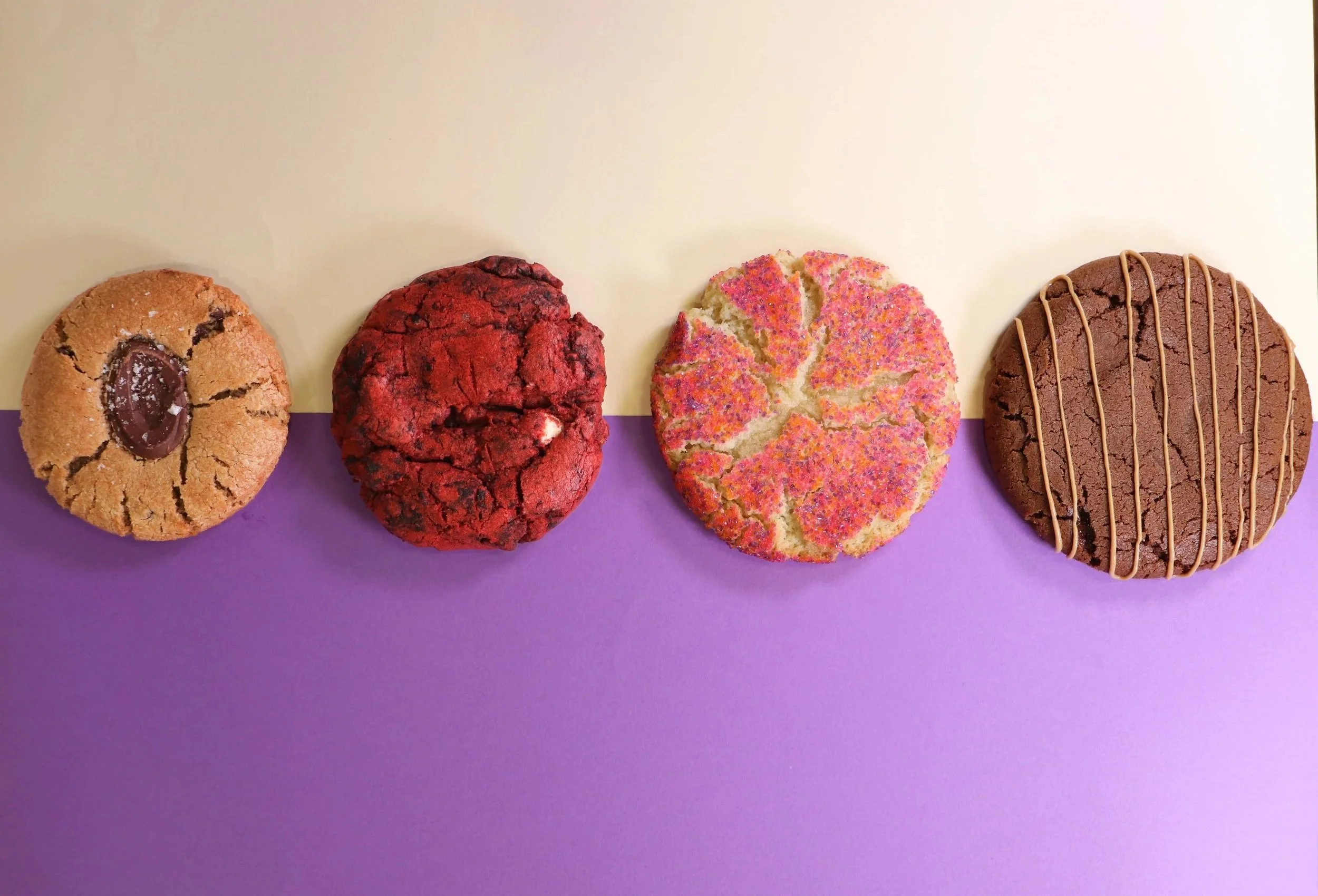 Four cookies arranged in a row on a surface with a purple and cream background. The first cookie is round with a chocolate center and cracks. The second is a large red cookie with a rough surface and chocolate chunks. The third is a round with pink and orange sprinkles on a light-colored base. The fourth is a round chocolate cookie with a drizzle of caramel.