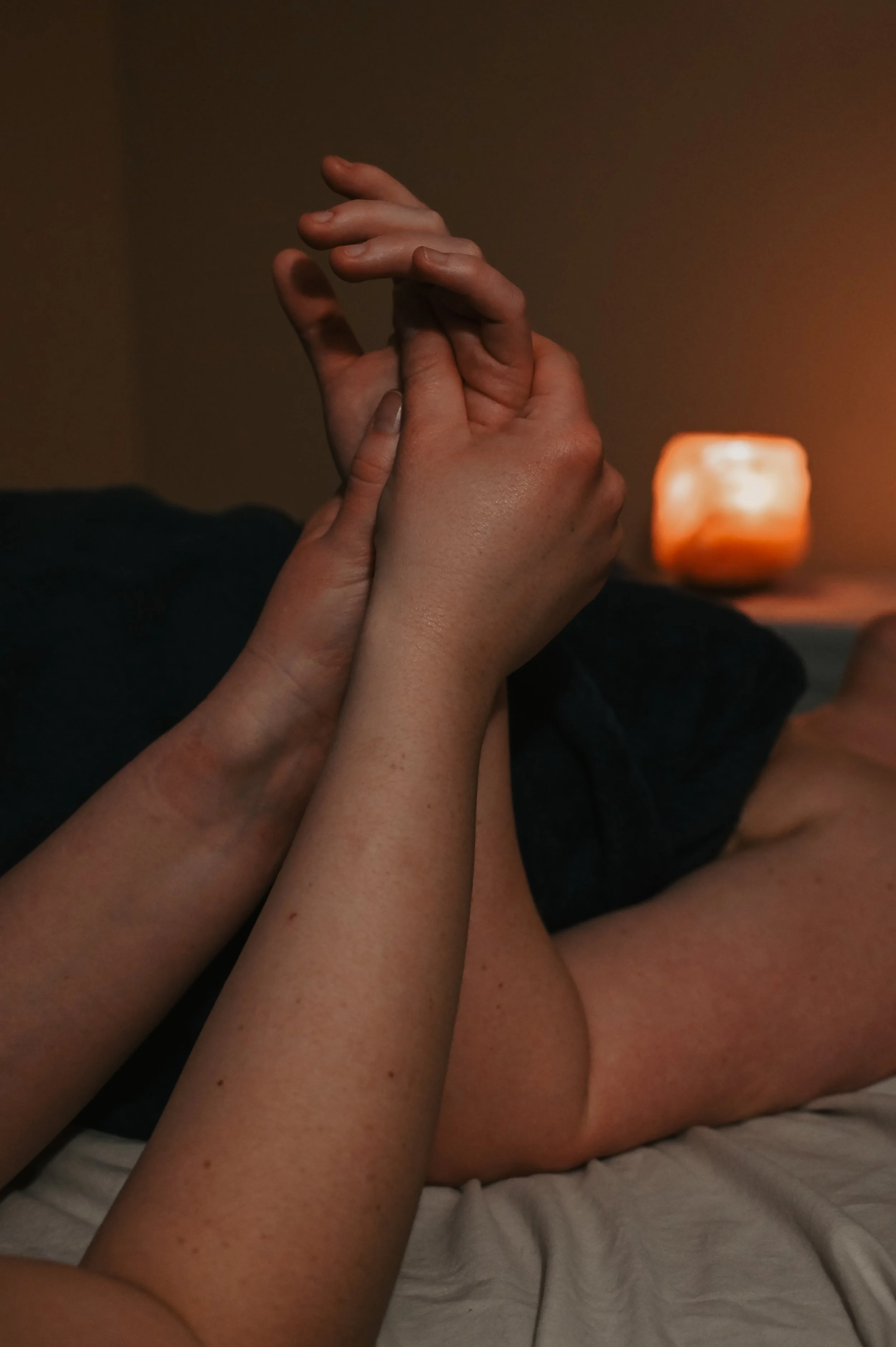 Two people are holding hands in a gentle and intertwined manner, with a soft, warm lamp glowing in the background.