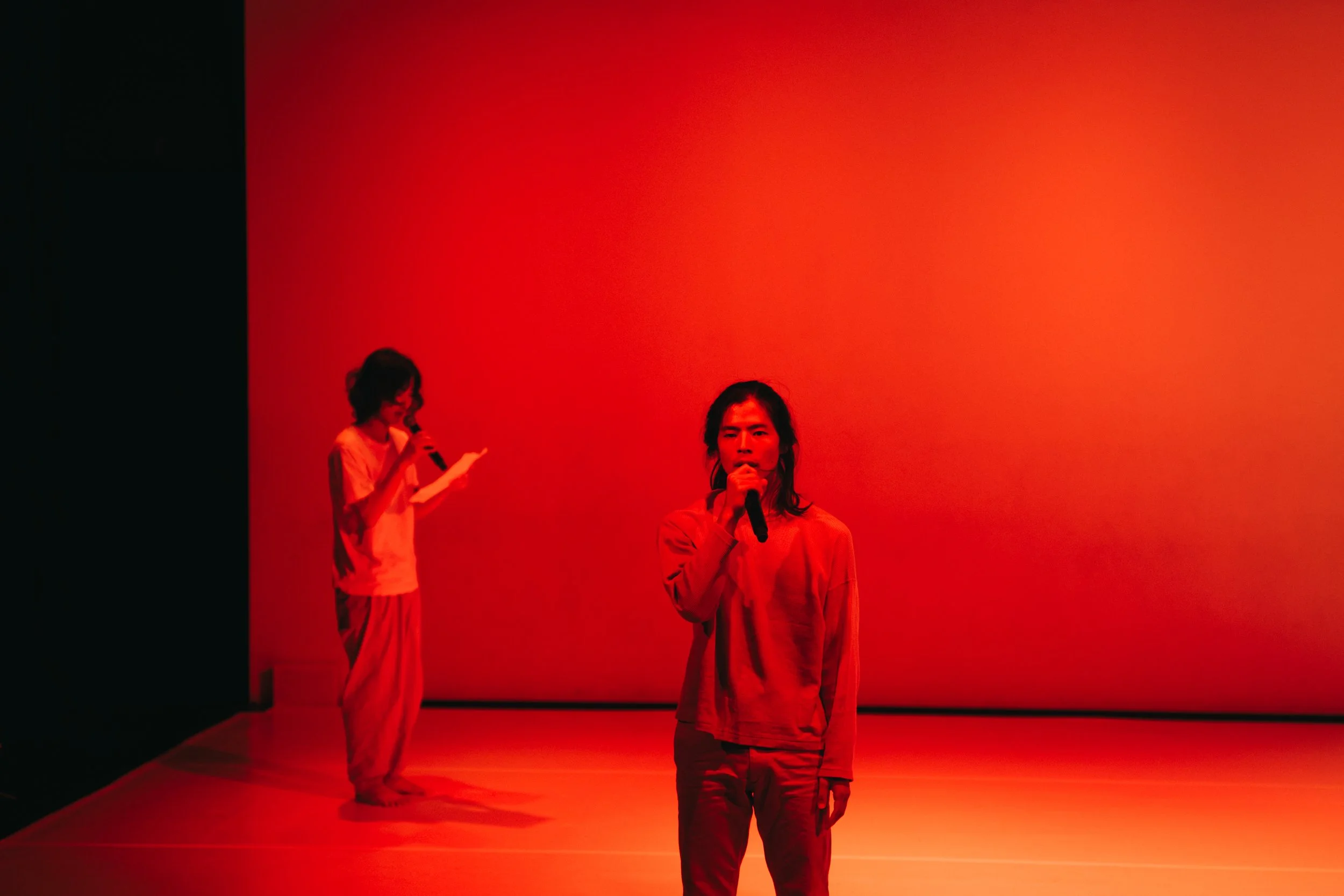 Endo Ryonosuke speaking into a microphone while Asahi Amano reads in Total Beings | Burning Hearts.