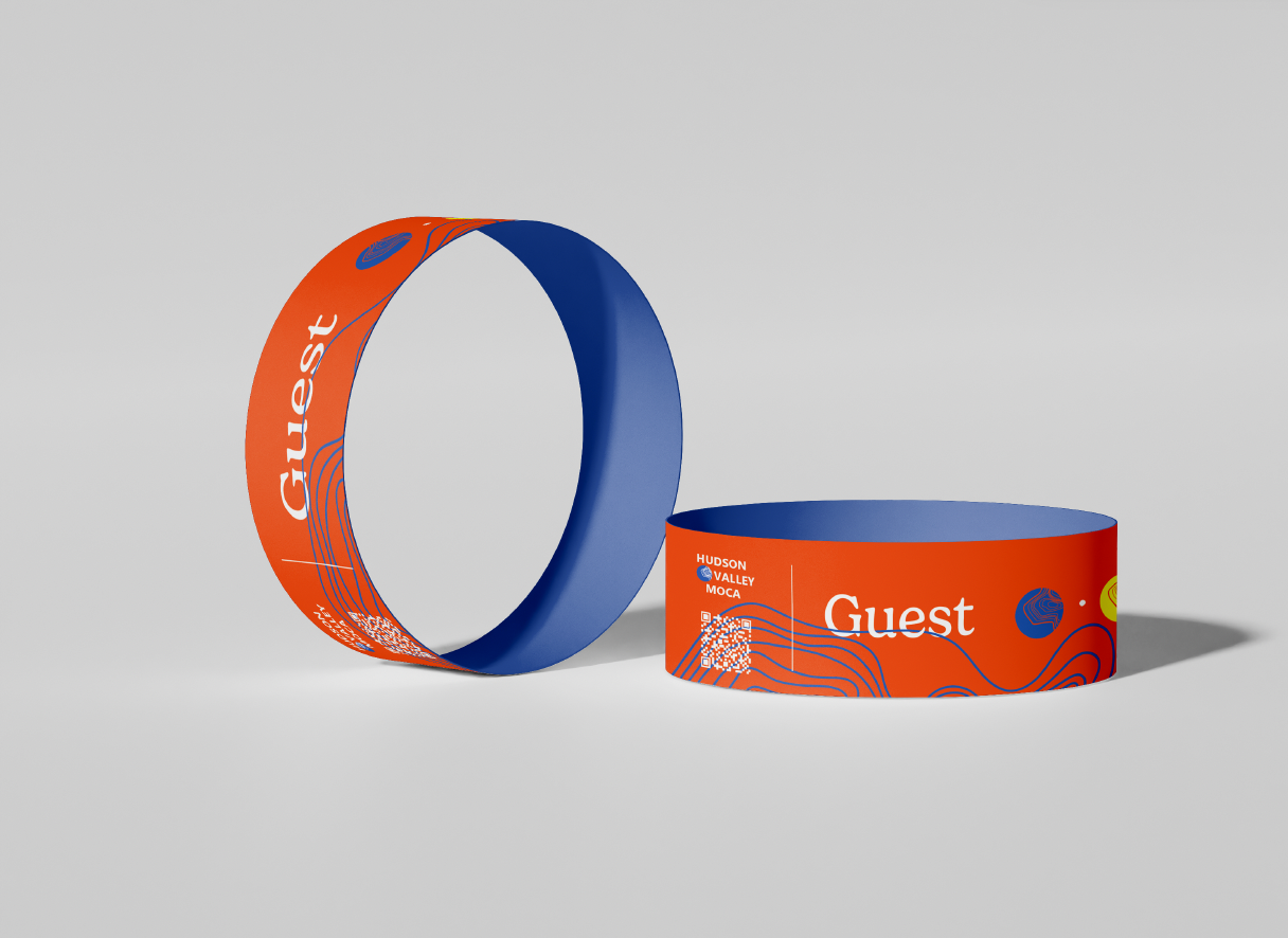 Two orange and blue wristbands with the word "Guest" printed on them, one lying flat and the other standing upright. The wristbands feature design elements including lines, a QR code, and text indicating event locations like Hudson Valley and MOCA.