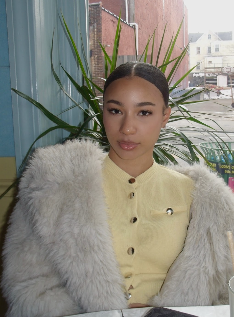 A young woman sitting indoors, wearing a yellow button-up top and a cream-colored fur coat, with her dark hair slicked back. Behind her are large green plants and a window showing a city street and buildings outside.