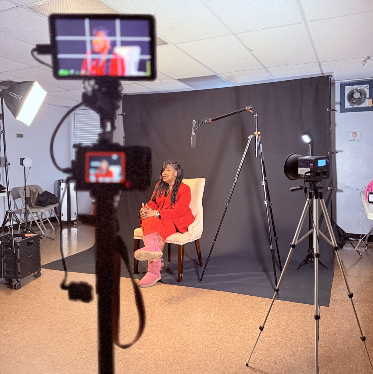 A woman in a red outfit sitting on a beige chair on a black backdrop in a photography studio, with filming equipment, lighting, and cameras around her.