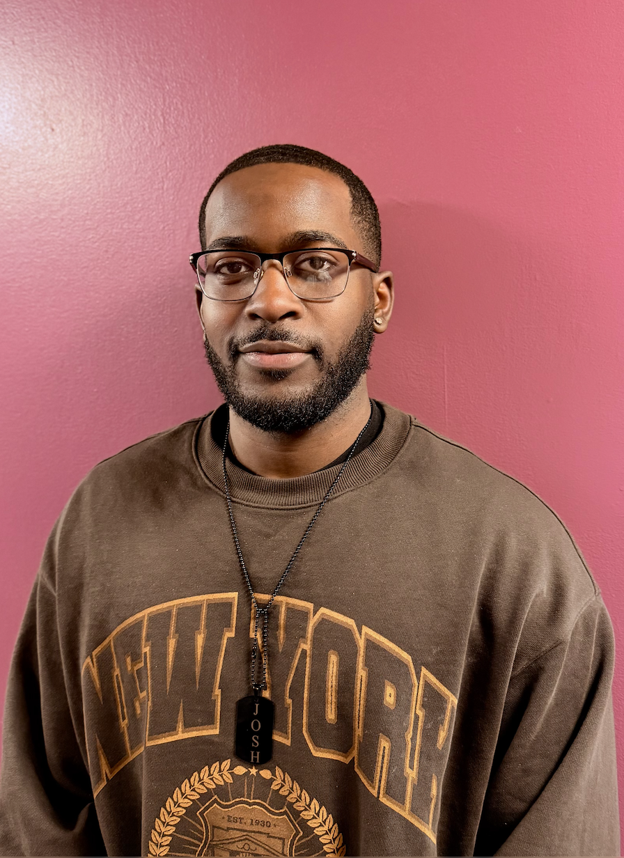 Young man with glasses, beard, and earrings, wearing a New York sweatshirt and a necklace with a pendant that has the name 'Josh,' standing against a pink wall.