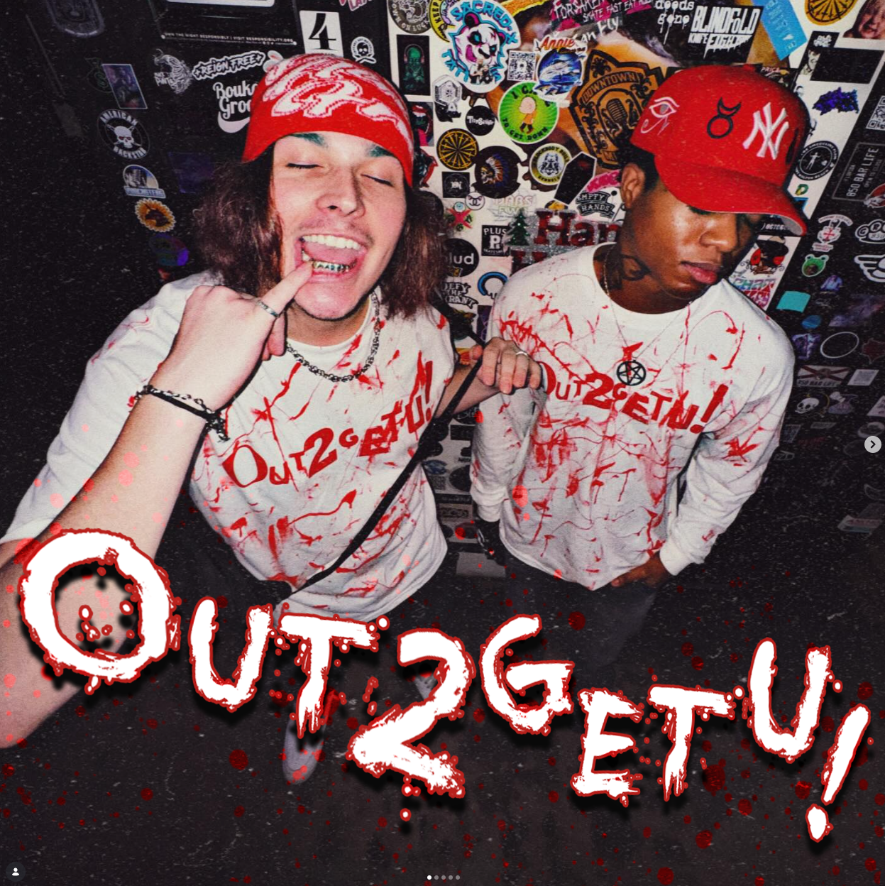 Two young men in a room decorated with stickers and posters, one smiling and showing his teeth, with a red bandana on his head and chain necklaces, the other looking down with a red cap and tattoos. They are wearing white shirts with red splatter designs. Red text and splatter graphics overlay the image, reading 'OUT2GET U!'.