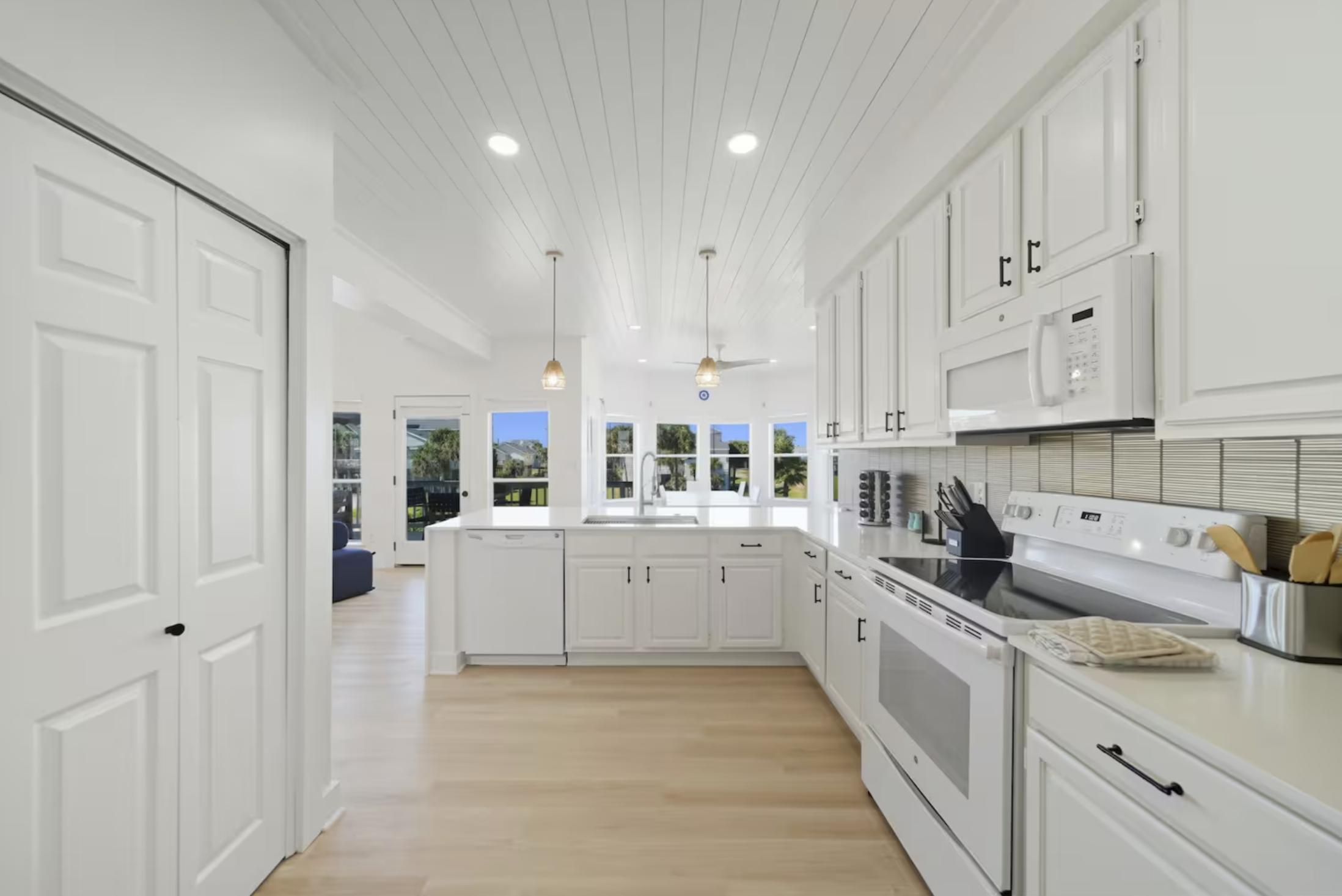 Bright, white kitchen with modern appliances, light wood flooring, and large windows in the background showing outdoor scenery.