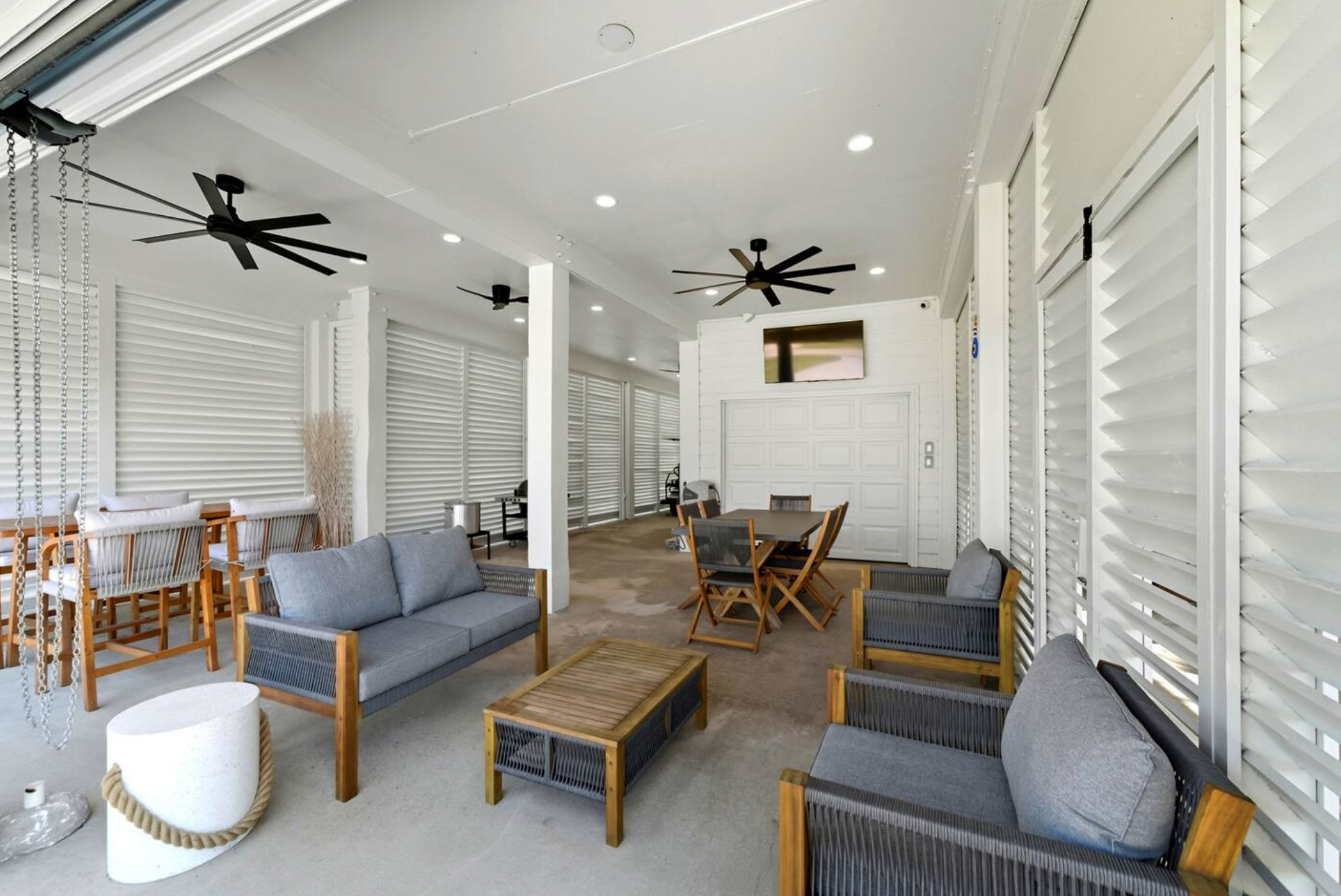 Bright modern lounge with gray couches, wooden chairs, ceiling fans, white walls, and white louvered windows.