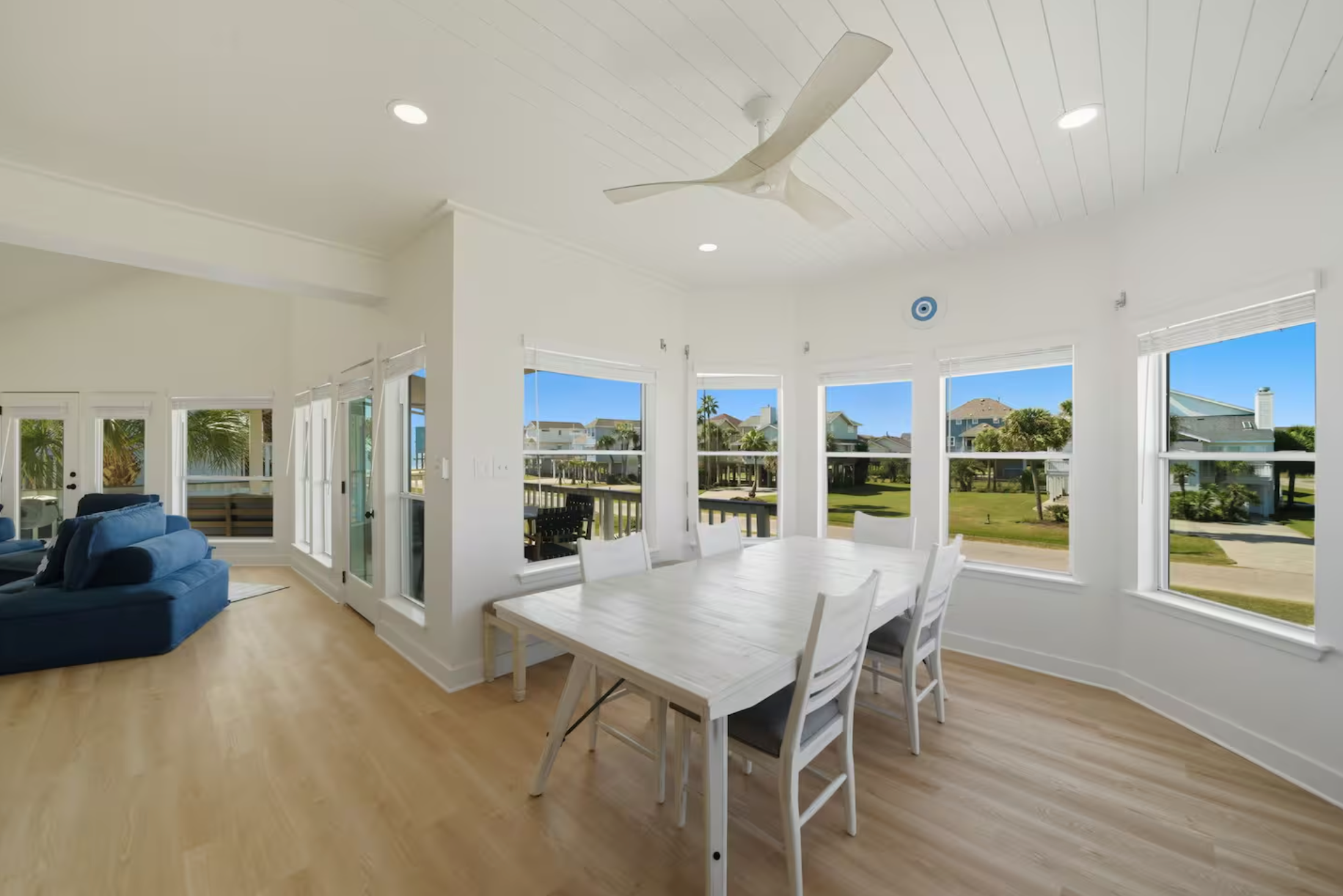 Bright, airy sunroom with large windows, white walls, wooden floor, white dining table with six chairs, ceiling fan, and visible outdoor greenery.