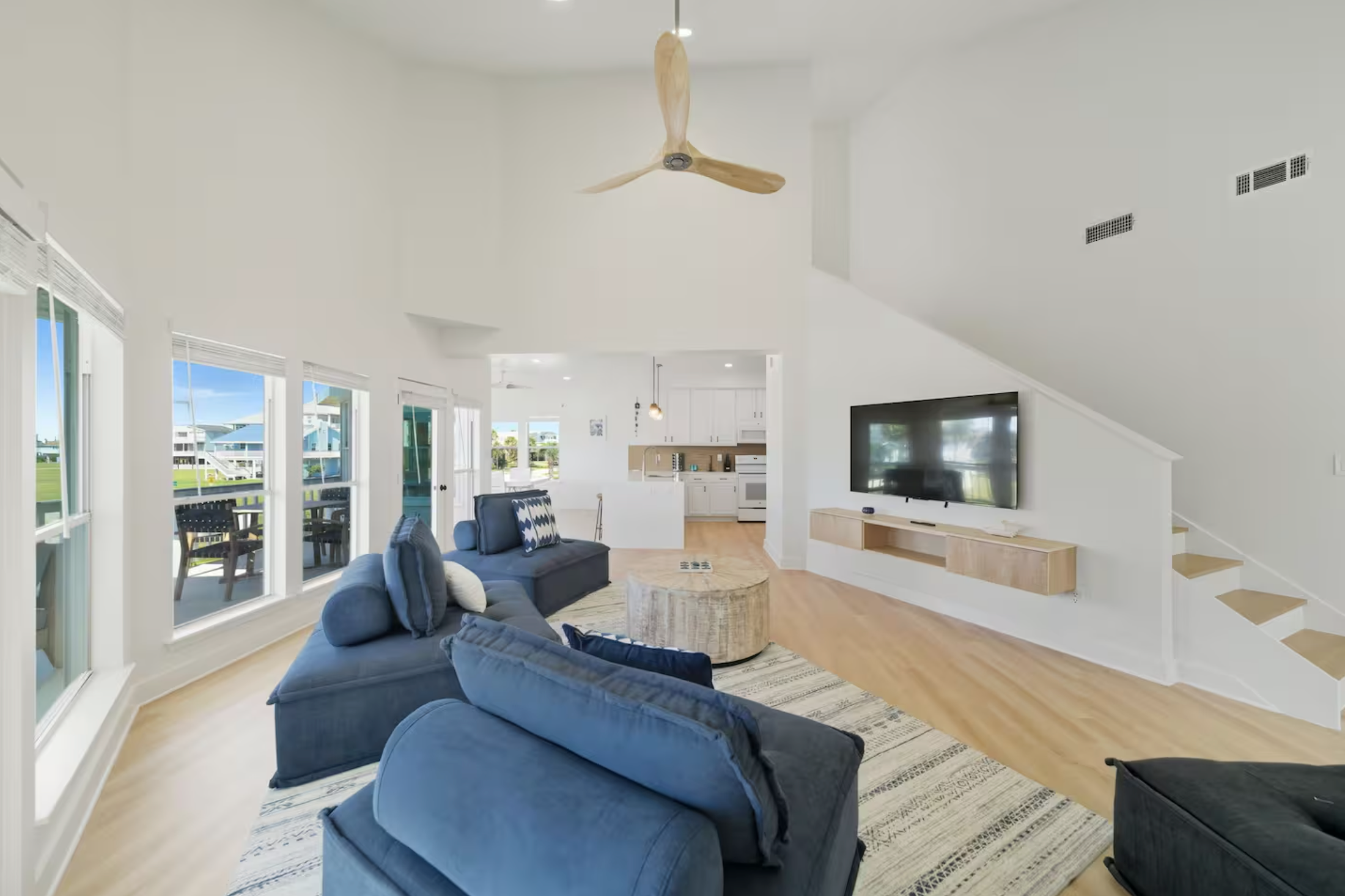 Living room with blue sofas, a round coffee table, wall-mounted TV, large windows, and a view of a porch with outdoor furniture.