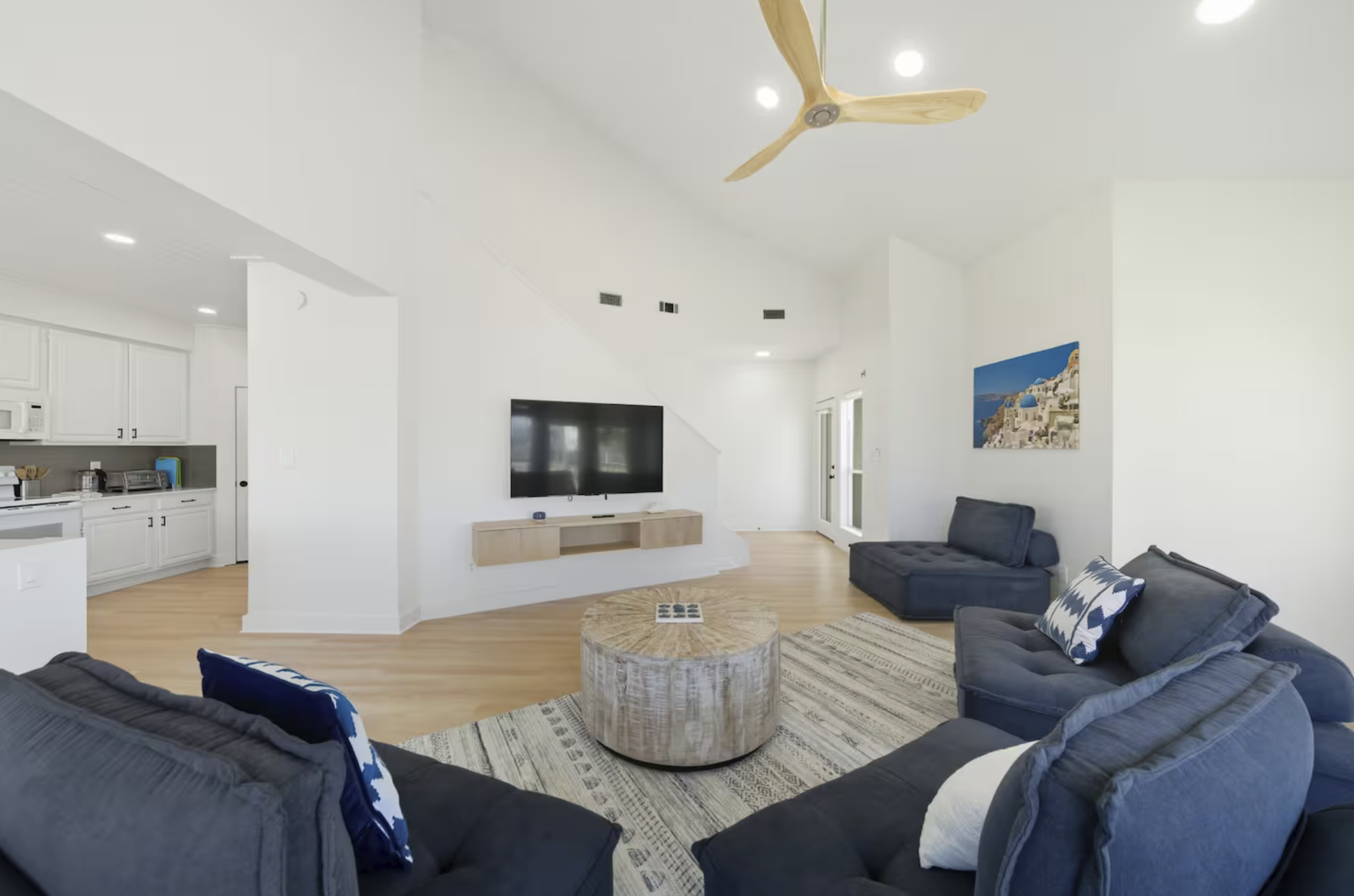Living room with dark gray sofas, a round wooden coffee table, a large flat-screen TV on the white wall, a painting of a coastal scene, and a ceiling fan with wooden blades.