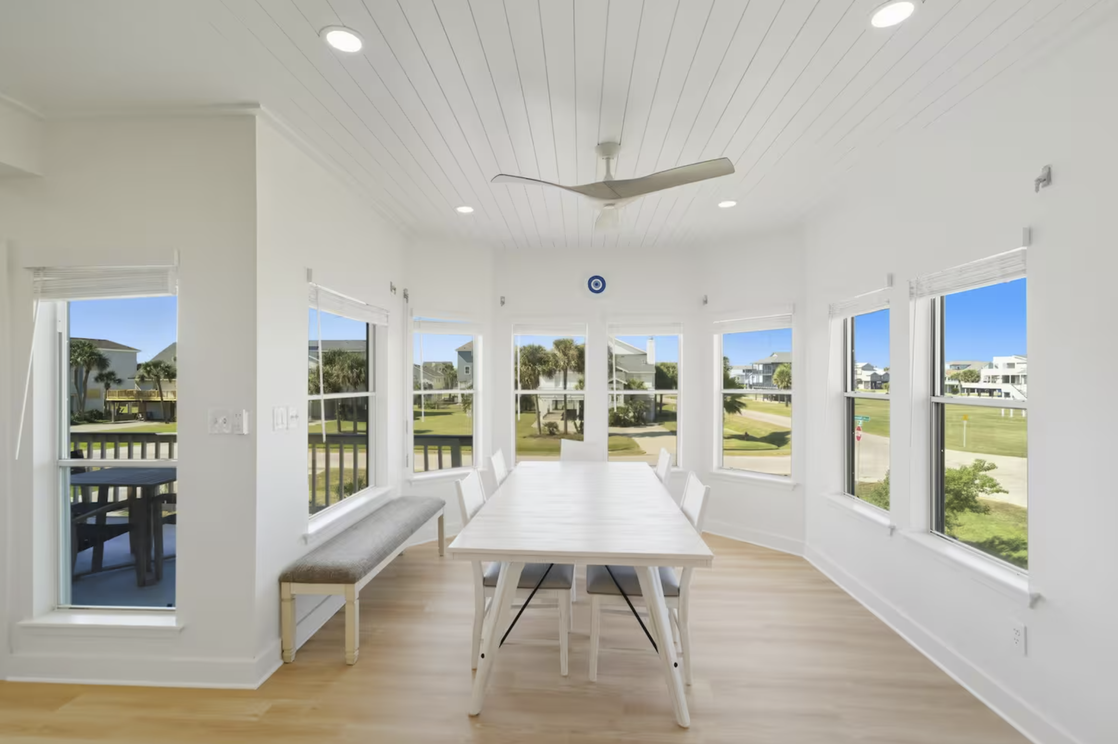 Bright dining room with large windows, white walls, light wooden floor, white table, six white chairs, ceiling fan, and view of outdoor greenery and houses.