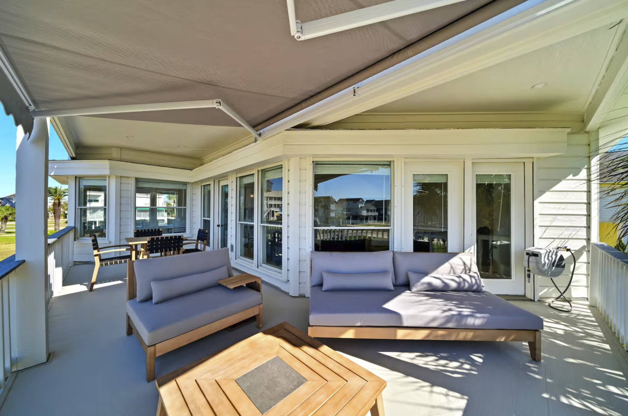 Outdoor covered balcony with gray cushioned seating, a coffee table, and a dining area with wooden chairs, overlooking neighboring houses and palm trees.